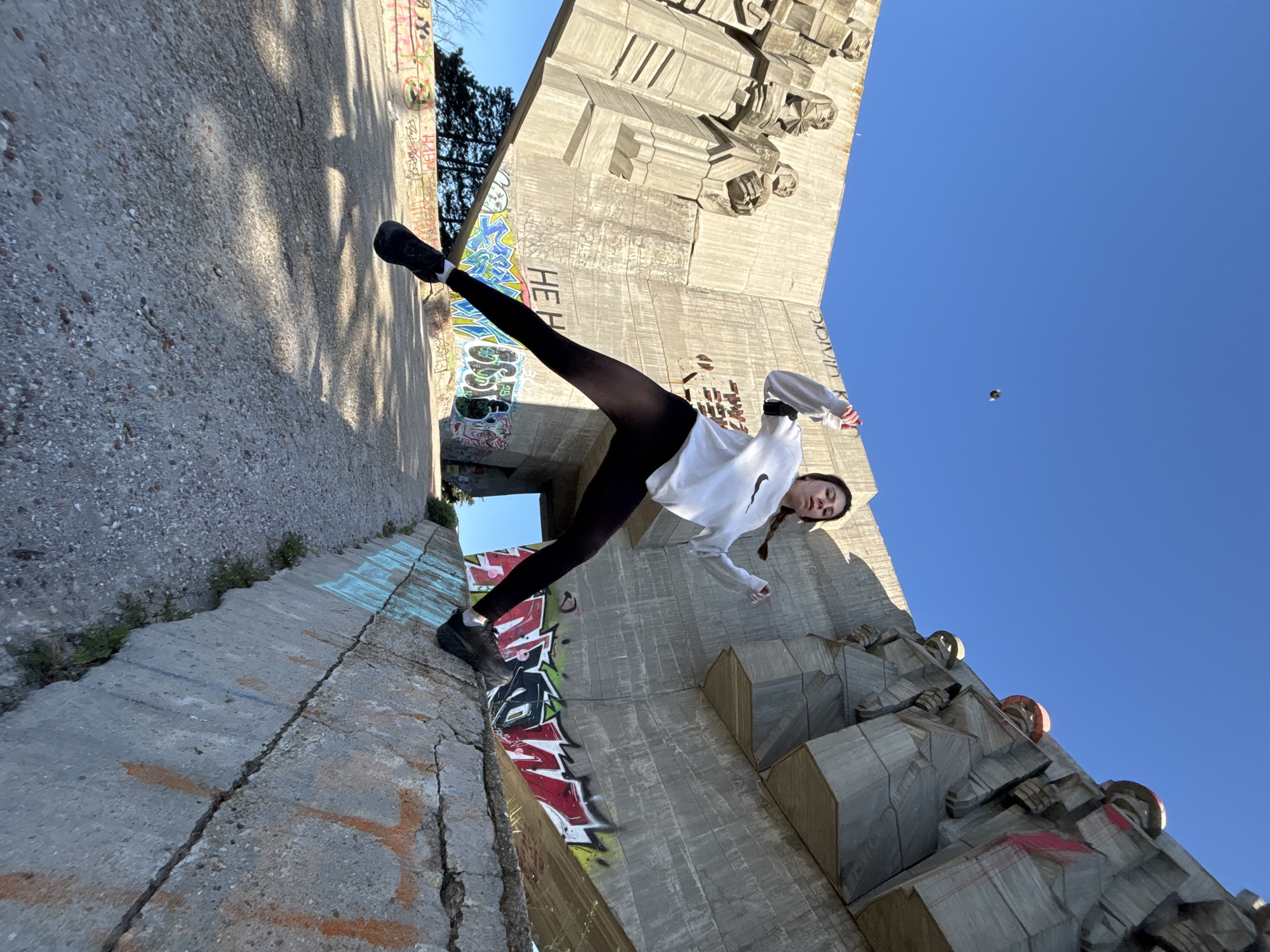 A young woman is captured in a dynamic, almost acrobatic pose against the backdrop of a monumental brutalist concrete structure under a bright, clear blue sky. The photograph is taken from a low angle and is rotated counter-clockwise, creating a striking and disorienting perspective.
The woman, appearing to be in her late teens or early twenties, has dark hair styled in two braids. She wears a white long-sleeved top with a dark logo visible on the chest (resembling a Nike swoosh), dark leggings, and dark athletic shoes. Her right leg is extended high, with her foot planted on a concrete ledge, while her body leans back and to the right, suggesting a moment of suspension or balance. A slight smile plays on her lips, conveying a sense of enjoyment or confidence.
The surrounding environment consists of massive concrete architecture, characterized by angular, block-like forms and sculptural reliefs. These structures are extensively covered in vibrant graffiti, featuring various tags and stylized letters in colors such as blue, green, red, black, yellow, and white. Portions of graffiti, including visible letters like "HEH," are present on the concrete wall behind the woman's extended leg. The ground and surrounding surfaces are also concrete, exhibiting cracks and additional graffiti.
The scene is brightly lit by natural daylight, indicating a sunny day, likely mid-morning or afternoon, with strong shadows visible on some of the concrete surfaces. The monument's imposing presence, combined with the woman's agile pose and the widespread graffiti, creates a sense of urban exploration or parkour within a public space. The location is identified as Varna, Bulgaria, suggesting the monument is a significant local landmark.