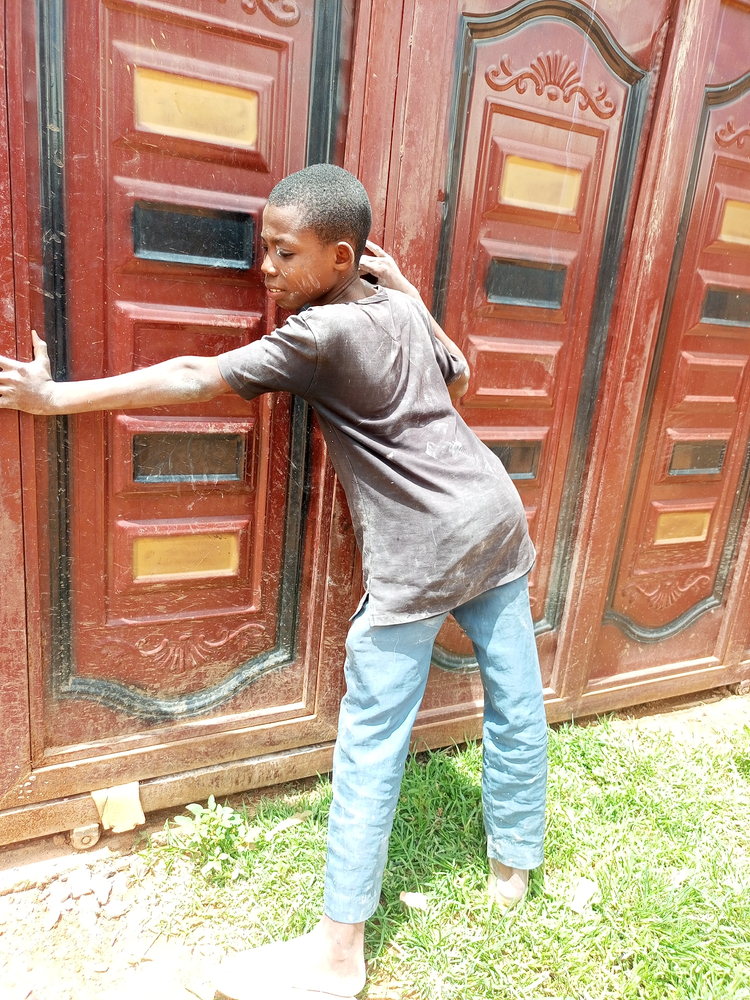 A young boy, appearing to be of African descent, stands with his left arm extended and pressing against a large, weathered, reddish-brown metal gate. The gate is segmented into panels, some of which have raised decorative elements. Visible on the gate are rectangular insets, one colored yellow and others appearing dark or black. The boy wears a dark, short-sleeved t-shirt and light blue trousers. His head is turned to his right, and his facial expression is not clearly discernible due to the angle. He is barefoot, with his feet on green grass and a patch of dirt or gravel. The environment suggests an outdoor setting, possibly in a yard or alleyway within the city of Tempure, Nigeria. The lighting indicates it is daytime, with sunlight casting shadows.