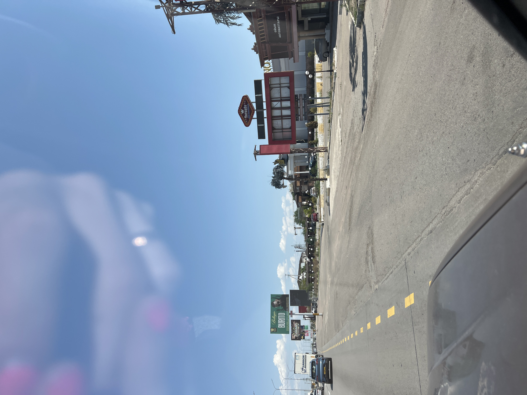 A wide asphalt road with a double yellow line and dashes separates two lanes of traffic, under a clear blue sky with scattered clouds. On the left side, a large white truck with the text "SARRION" is in the right lane, with a dark blue car in the left lane. Several billboards are visible on the left side of the road. The largest billboard features an image of a man with the text "Bellagio BURITO" and a phone number. Another billboard further down displays "8 700 000 000". On the right side of the road, a complex of buildings with red and beige facades is visible. A large red sign with "Tornado" written in a star-shaped logo is prominent. Another building has text in Cyrillic, likely "OYIN AVTOMATLARY" which translates to "game machines." The scene suggests a commercial area or entertainment complex.