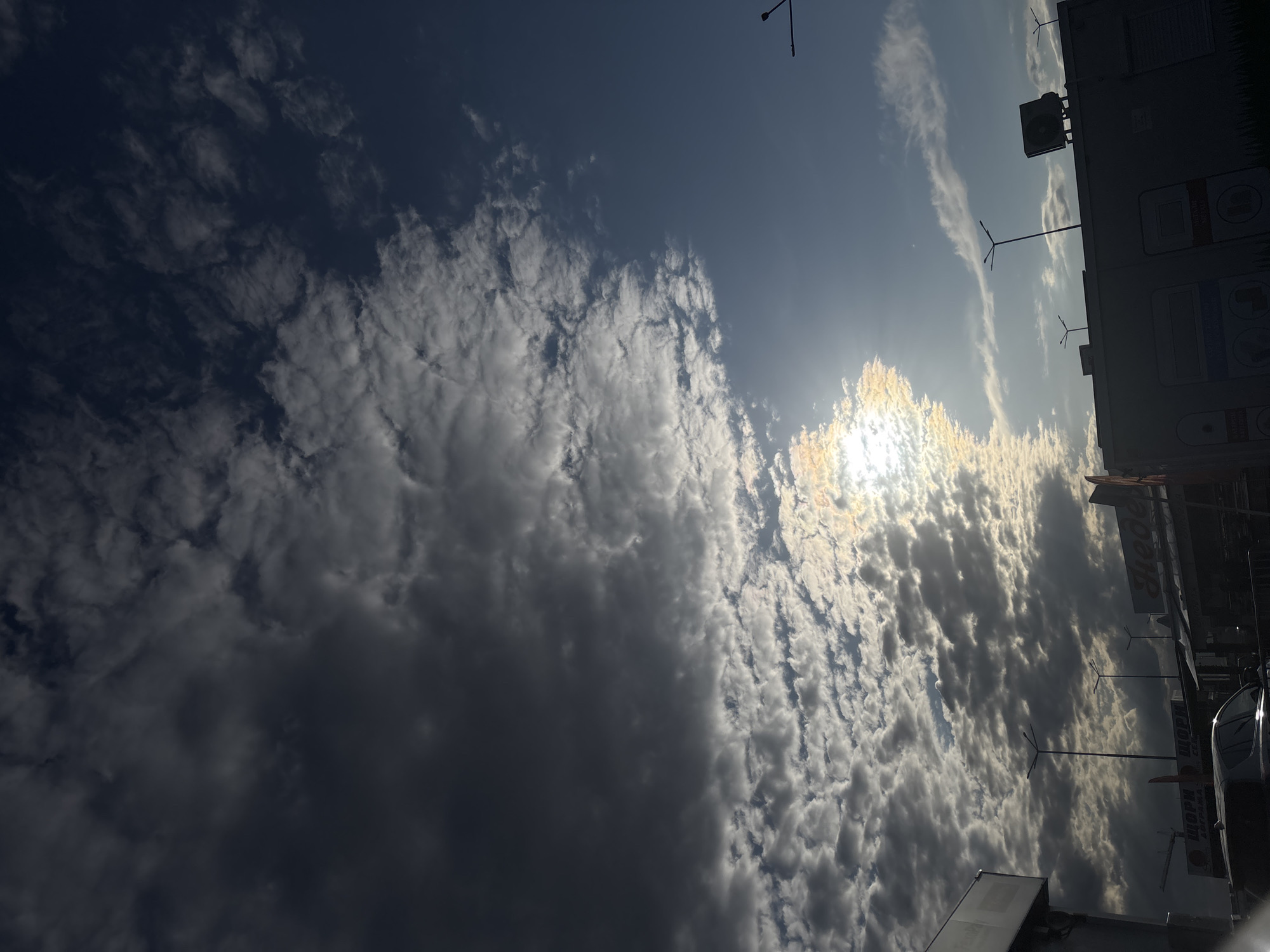The image shows a view of the sky above Sofia, Bulgaria. The sky is partly cloudy, with the sun shining through a break in the clouds. Some buildings and street lights can be seen in the background. The image appears to have been taken at an angle, tilted slightly.