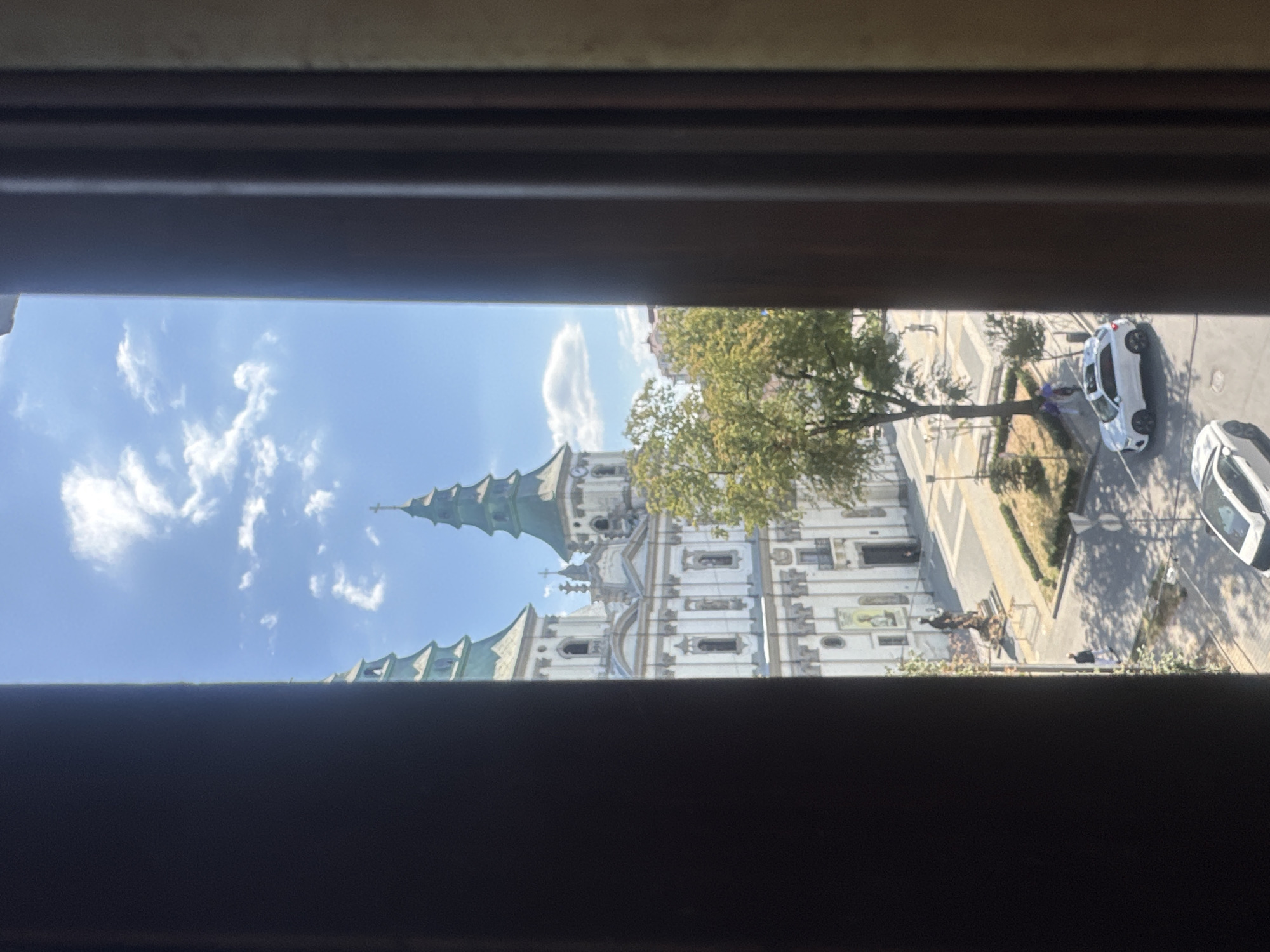 The image shows a view of the Church of the Immaculate Conception of the Blessed Virgin Mary in Ternopil, Ukraine, through a window frame. The church features two prominent green spires and a white facade with intricate architectural details. In front of the church, there is a paved square with a statue in the center, surrounded by trees and a street. Several people are visible in the square and on the street, some walking and some standing. Two white cars are parked on the street, and another car is driving. The sky is blue with scattered white clouds, indicating a clear, sunny day. The overall scene depicts a peaceful urban environment with a significant religious landmark.