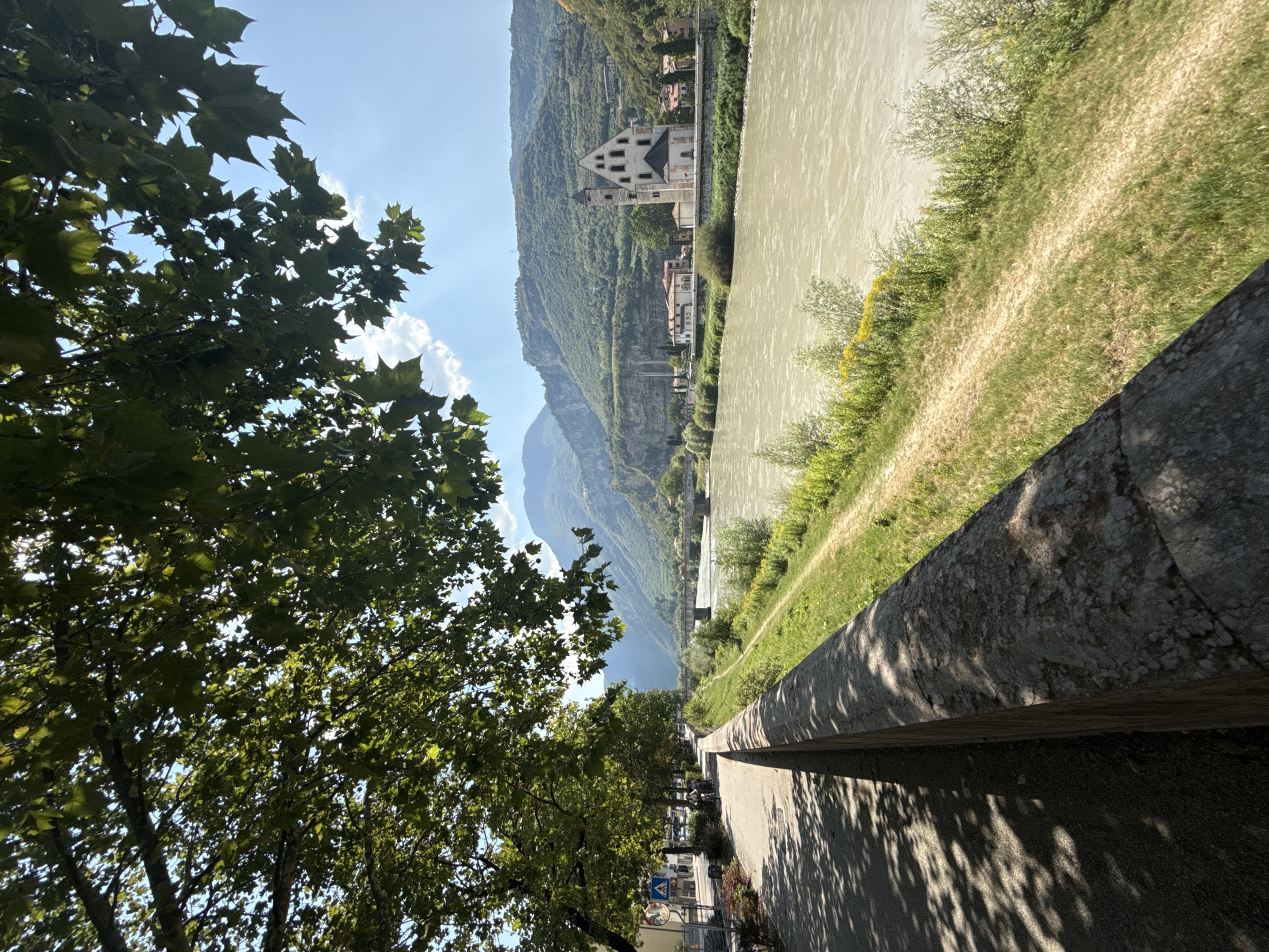 The image depicts a scenic view of the Adige River in Trento, Italy, from a riverside promenade. A stone wall runs along the left side of the frame, separating the promenade from a grassy embankment. The promenade is paved and shaded by the leaves of a large tree in the foreground. On the left side of the promenade, a street with some parked cars and pedestrians is visible. Across the river, a church with a prominent bell tower stands against a backdrop of lush green hills and mountains. A bridge spans the river in the distance. The sky is blue with a few scattered clouds. The overall impression is one of a peaceful and picturesque urban landscape.