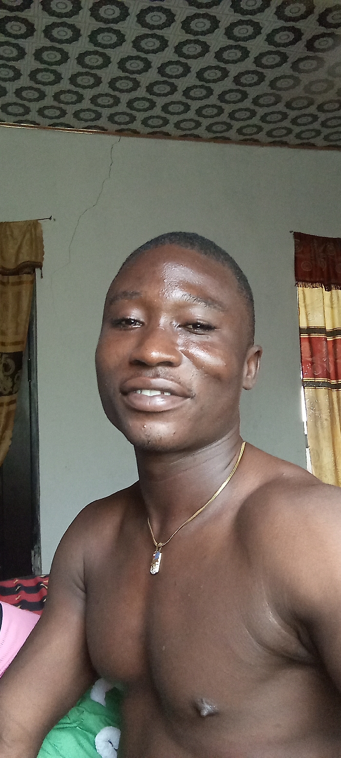 An image taken indoors in Dzodze, Ghana, depicts a shirtless male subject of dark complexion, facing forward and slightly to the right. He has short dark hair and is smiling, revealing upper and lower teeth. A gold-colored chain necklace with a rectangular, multi-colored pendant rests on his chest. His skin exhibits a sheen. The background features a patterned ceiling with dark, circular motifs on a lighter surface. A vertical crack is visible on the light-colored wall extending downwards from the ceiling line on the left side of the frame. Two curtains are partially visible: one on the far left, brown/tan with dark vertical stripes, and another on the far right, reddish-brown and tan/gold striped with a fringed lower edge.