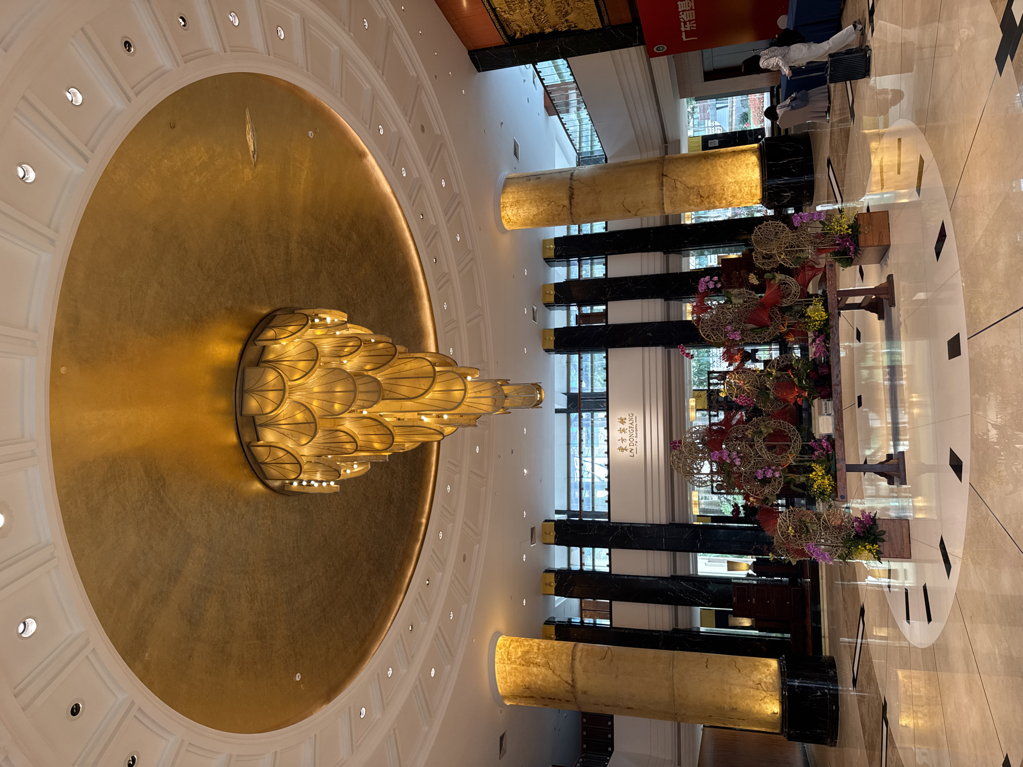 The image shows the interior of what appears to be a hotel or luxury building in the Yuexiu District of China. Dominating the view is a large, ornate chandelier hanging from a circular gold-colored ceiling. The ceiling is surrounded by a ring of recessed lights set into a white, coffered structure. Below, the floor is polished and reflective, with geometric patterns. Columns, some with gold-colored accents and some with dark marble bases, punctuate the space, dividing it into sections. Tables decorated with floral arrangements are present, adding to the upscale ambiance. To the right side of the image, people can be seen standing with luggage, suggesting a hotel lobby setting. Signage with Chinese characters is visible above the columns. Overall, the scene conveys a sense of luxury and grandeur.