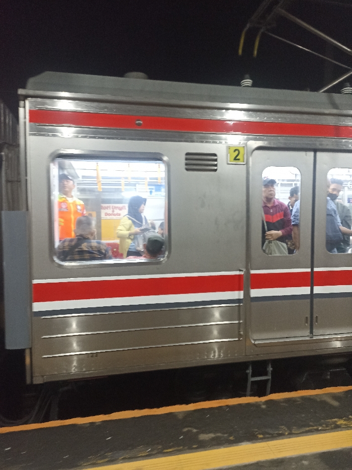 The image shows a train car at the Bojong Gede train station in Indonesia, located at latitude -6.494 and longitude 106.795. The train car is painted grey with red and white stripes. The car is numbered '2' and has a large window revealing a view of the train's interior, where a man in a yellow uniform stands with his back to the camera, facing a woman in a hijab. Another woman in a hijab stands with her back to the camera, talking to a man in a red plaid shirt. In the background, a man wearing a blue shirt and a man wearing a black shirt can be seen. The scene takes place at night and the only source of light appears to come from within the train car itself. A person appears to be standing on the platform. The image is taken from a low angle, with the camera pointing slightly upwards. The track and platform are dark and only partially lit, with a bright yellow painted line bordering the platform. The track and platform appear empty.