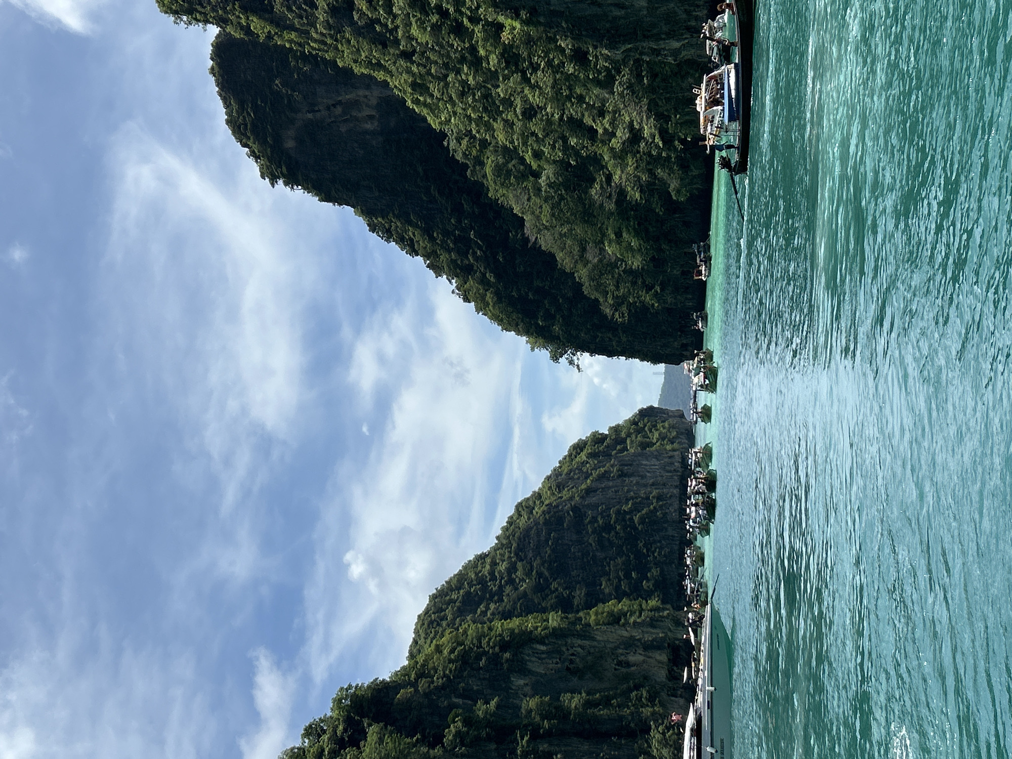 The image depicts a tropical seascape with towering, verdant cliffs framing a body of turquoise water. Numerous boats, including longtail boats and a larger white vessel, are scattered across the water, some appearing to be anchored and others in motion. The cliffs are covered in dense vegetation, suggesting a lush, natural environment. The sky above is partly cloudy with a mix of blue sky and white clouds, indicating fair weather. This scene is characteristic of the iconic limestone karst landscape found in and around Ao Nang, Thailand, known for its stunning bays and islands.