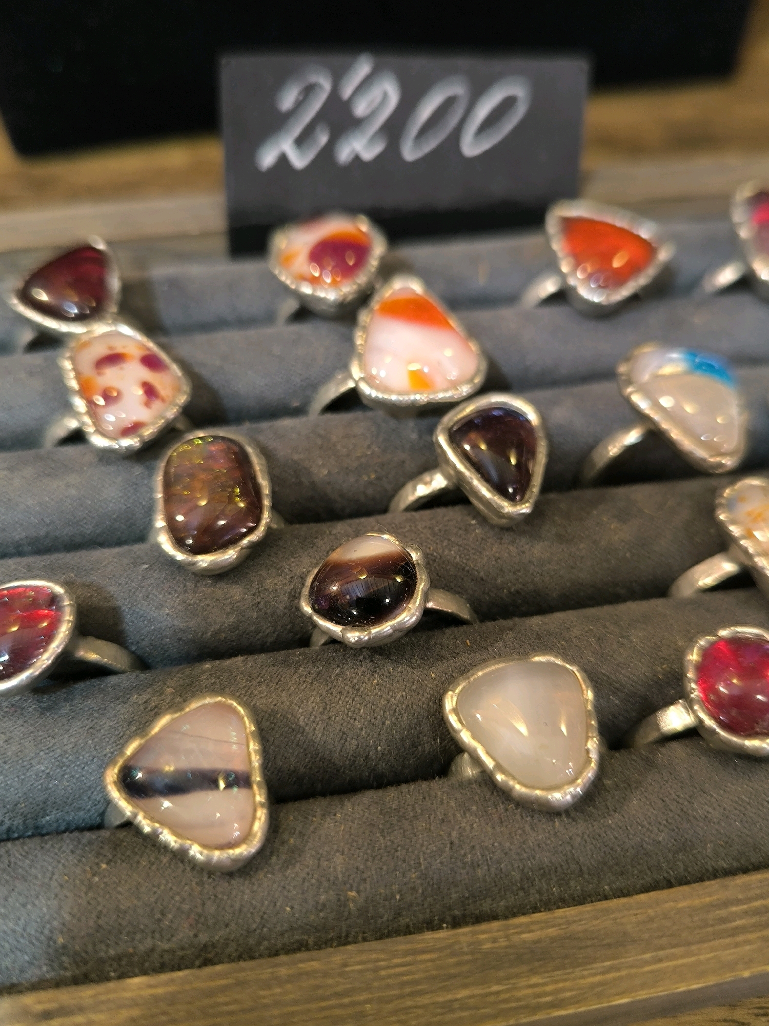A close-up, eye-level shot showcases an assortment of rings displayed on a dark gray, fabric-covered surface. The rings, each featuring a unique, polished stone or colorful glass cabochon set in a silver-toned metal band, are arranged in rows. The stones vary in shape and color, including deep reds, oranges, purples, whites, and blues, with some appearing translucent or opalescent. In the upper background, a black sign with white, stylized lettering reads "2200," presumably indicating a price. The lighting is soft, highlighting the reflections and textures of the jewelry. The overall setting suggests a retail display, possibly in a jewelry store or market stall. There are no people or animals visible in the image.