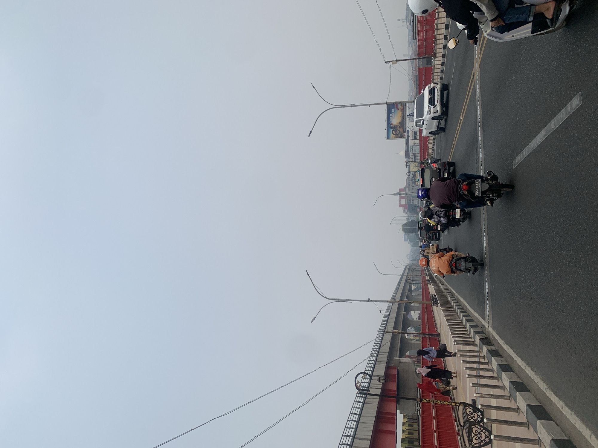The image depicts a daytime scene on a bridge in Palembang, Indonesia, likely the Ampera Bridge, under an overcast sky. The bridge carries traffic, with several motorcycles and a white SUV visible. A pedestrian walkway runs alongside the road, separated by a red railing. Several people are walking on the sidewalk, some wearing headscarves. The environment appears to be urban, with buildings and streetlights visible in the distance. The overall impression is one of moderate activity on a bridge connecting different parts of the city.