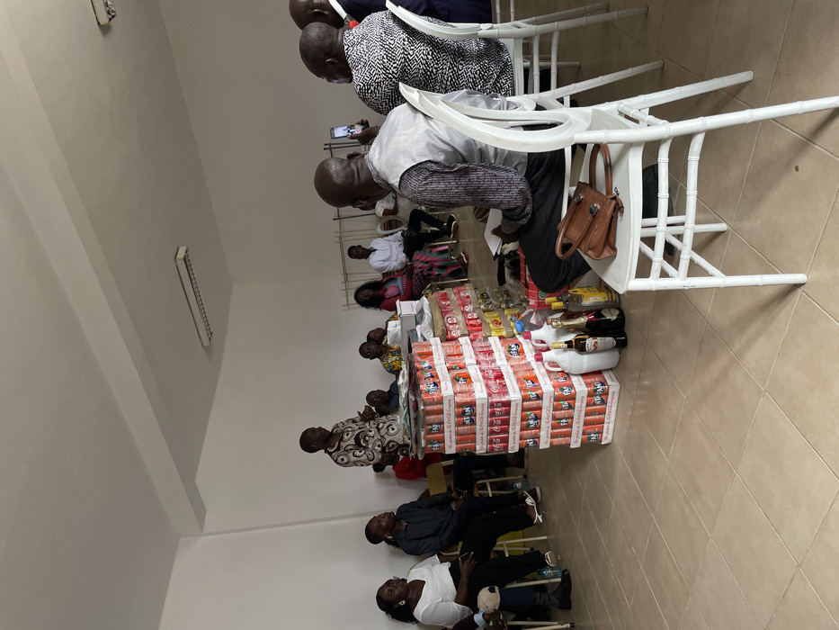 This image captures an indoor gathering in what appears to be a community hall or event space in Luanda, Angola. The room features light-colored walls and a tiled floor.
Numerous people, predominantly adults, are seated on white plastic chairs arranged in rows, facing towards the right side of the frame. The perspective is from the front-left of the assembled group. In the foreground and middle ground, individuals are seen observing or engaged with their personal items, such as mobile phones. One person in a light-colored shirt and dark trousers has a brown leather handbag hanging from the back of their chair. Another individual nearby appears to be holding papers.
The central focus of the scene is a substantial display of various beverages arranged on what seems to be a table or platform. The base of this display is formed by stacked boxes of soft drinks, with "FANTA" and "Coca-Cola" branding prominently visible on the cans and cartons. The text "THE COCA-COLA COMPANY" is also legible on some boxes. On top of these stacked soft drink cases, an assortment of bottled alcoholic beverages, including what appear to be whiskey and wine, are meticulously arranged alongside several clear plastic jugs.
The event suggests a form of presentation, perhaps a ceremony involving gifts or donations, or a social gathering where these items are displayed for distribution. The lighting within the room is artificial, with a fluorescent light fixture visible on the upper part of the left wall. The attendees are dressed in a mix of casual and semi-formal attire, and their expressions appear calm and attentive.
