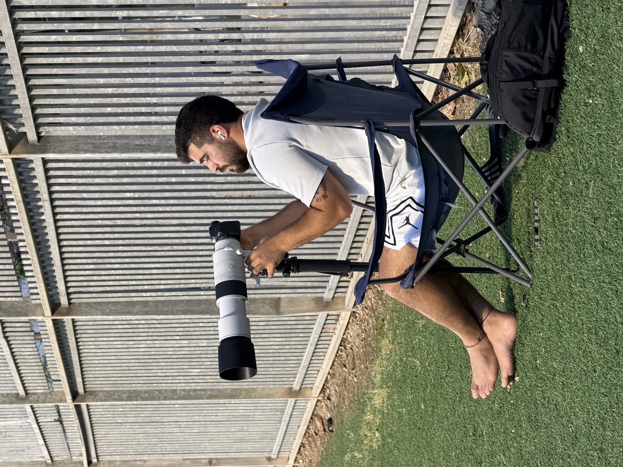 A man with a beard and short dark hair sits in a blue folding chair on a patch of green grass. He is wearing a light-colored short-sleeved shirt and white athletic shorts with a black logo. He holds a large white telephoto camera lens attached to a camera. The camera is mounted on a black monopod. A black backpack sits behind him on the grass. Behind him is a gray metal fence structure. The location is Tel-Aviv, Israel.