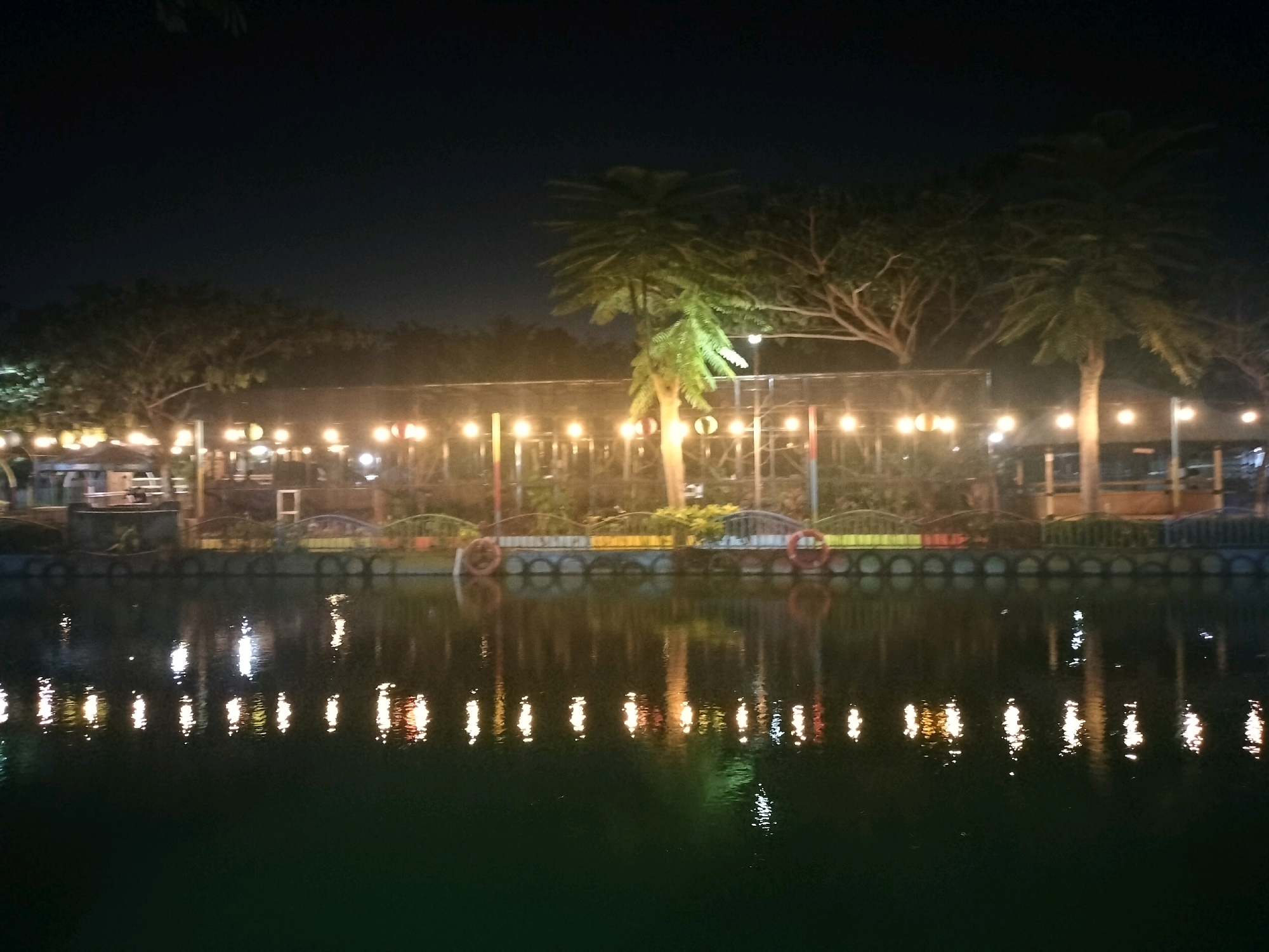 The image captures a nighttime scene at the Grand Opening of the Setu Citayam Park in Depok, Indonesia. The park, situated adjacent to a body of water, is illuminated by a string of warm-toned lights that hang above a decorative fence. This fence, composed of tire-like structures and colorful panels, borders the water. Two life preservers are visible, one on each side of the fence. In the background, several trees, including palm trees, are silhouetted against the night sky. The lights from the park are reflected in the dark water, creating a shimmering effect. The scene suggests a festive atmosphere, likely during an event or opening.