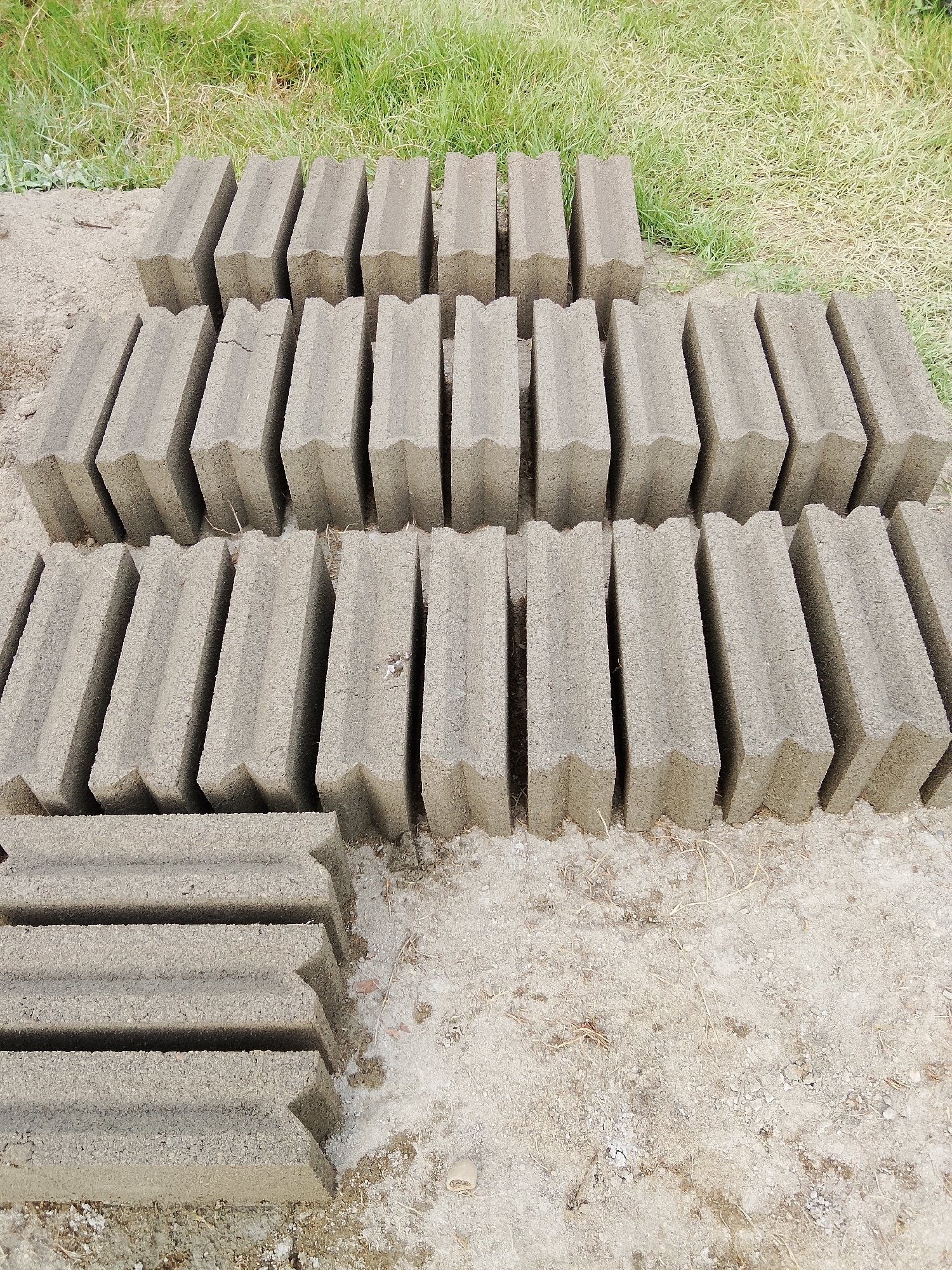 The image shows a collection of concrete blocks arranged in neat rows on a patch of dry soil and short grass. The blocks are rectangular with a distinctive interlocking shape, featuring a zig-zag pattern along one edge that allows them to fit together securely. They are a uniform light gray color, typical of concrete.
In the foreground, several rows of these blocks are stacked horizontally. Behind them, the blocks are arranged vertically, forming several distinct columns. The overall arrangement suggests they are either newly made and drying, or are being prepared for construction. The setting appears to be outdoors, with a background of green grass, hinting at a somewhat rural or undeveloped area. The ground is covered with a mixture of soil, small stones, and dry grass or straw, indicating a dry environment.
There is no visible text in the image. The lighting suggests it is daytime, with no strong shadows indicating it is either early morning or late afternoon. The weather appears to be dry. The image is taken from a slightly elevated perspective, looking down at the blocks. The scene is one of simple industry or preparation, with a focus on the utilitarian objects. The location context of Kruenglingka Barat, Indonesia, suggests a tropical or subtropical climate, which aligns with the presence of dry soil and grass.