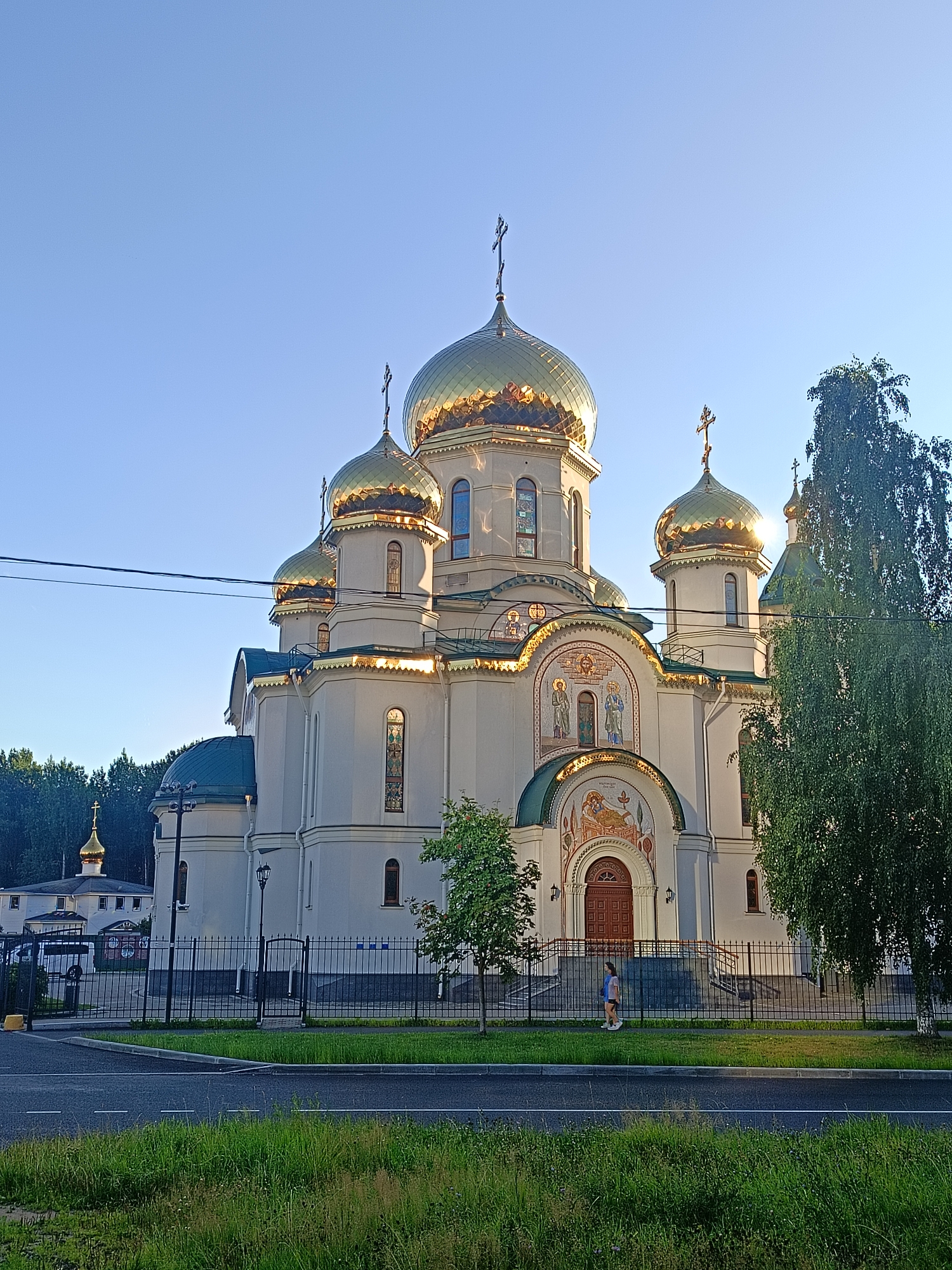 The image shows the Church of the Resurrection of Christ in Saint Petersburg, Russia. The church is a large, white building with multiple golden domes, characteristic of Russian Orthodox architecture. It is set against a clear blue sky, with lush green trees and grass surrounding it. A black metal fence separates the church grounds from a paved road and sidewalk. A single person, a young woman, is walking on the sidewalk in front of the church. The sun is shining, casting a warm glow on the building and its golden domes. In the background, a smaller building with a golden dome can be seen, suggesting it might be part of a monastic complex or another religious structure. Power lines are visible in the sky, indicating the presence of urban infrastructure. The overall impression is one of peaceful, majestic religious architecture bathed in sunlight.