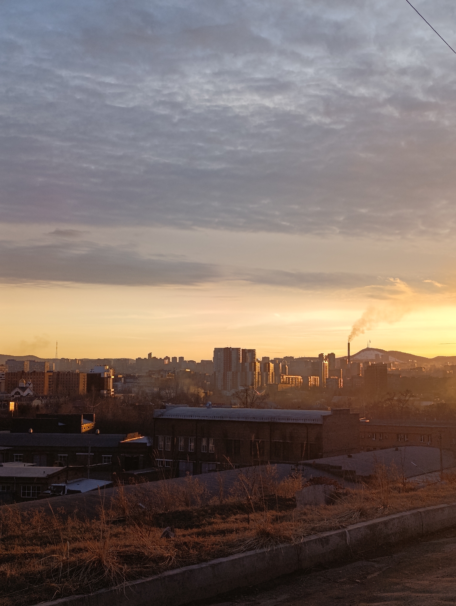 The image captures a cityscape at dawn or dusk, possibly in Krasnoyarsk, Russia, given the industrial landscape and architectural styles. The sky is a blend of grey clouds and orange hues from the rising or setting sun. In the midground, silhouetted buildings dominate the view. A smokestack emits a plume of smoke, suggesting industrial activity. The foreground features overgrown vegetation and a concrete barrier, suggesting a neglected or overlooked space within the city. The overall scene evokes a sense of urban decay juxtaposed with natural beauty.