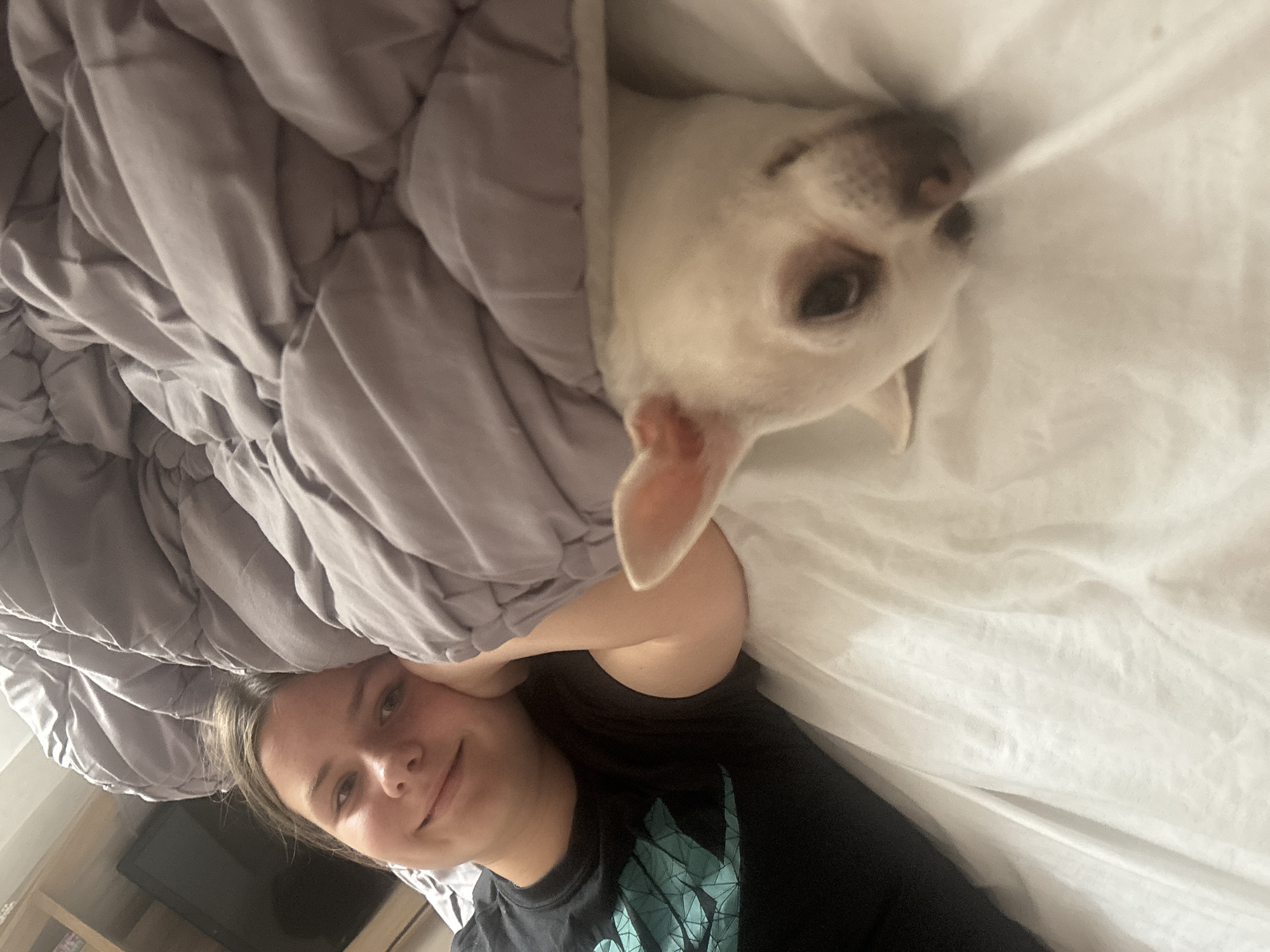 This image shows a young person lying in bed, with their head resting on their hand. A small white dog is peeking out from under the covers next to them. The room is lit by artificial light, and a shelf unit with a television is visible in the background. The scene appears to be indoors, possibly a bedroom. There is no information available to suggest this image is located in Shepetivka, Ukraine.