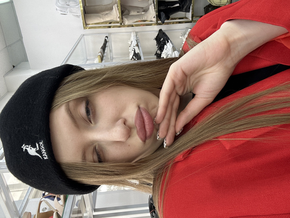 The image displays an adult female captured in a close-up, indoor self-portrait within what appears to be a retail establishment in Chaikovsky, Russia. The subject has long, light brown hair and wears a black beret with a white "KANGOL" logo and kangaroo emblem, along with a red blazer over a dark top. She exhibits a facial expression with her right eye partially closed and her lips pursed in a "kissy face." Her right hand, featuring fingernails with white bases and black polka dots, is positioned beneath her chin. The background reveals white walls and multiple display shelves containing various footwear. To the left, a clear, multi-tiered shelf holds sandals and sneakers. To the right, a gold-framed display shelf showcases several pairs of boots, including light-colored tall boots and black chunky boots. The precise location or name of the commercial establishment within Chaikovsky, Russia, cannot be determined from the visual information provided.