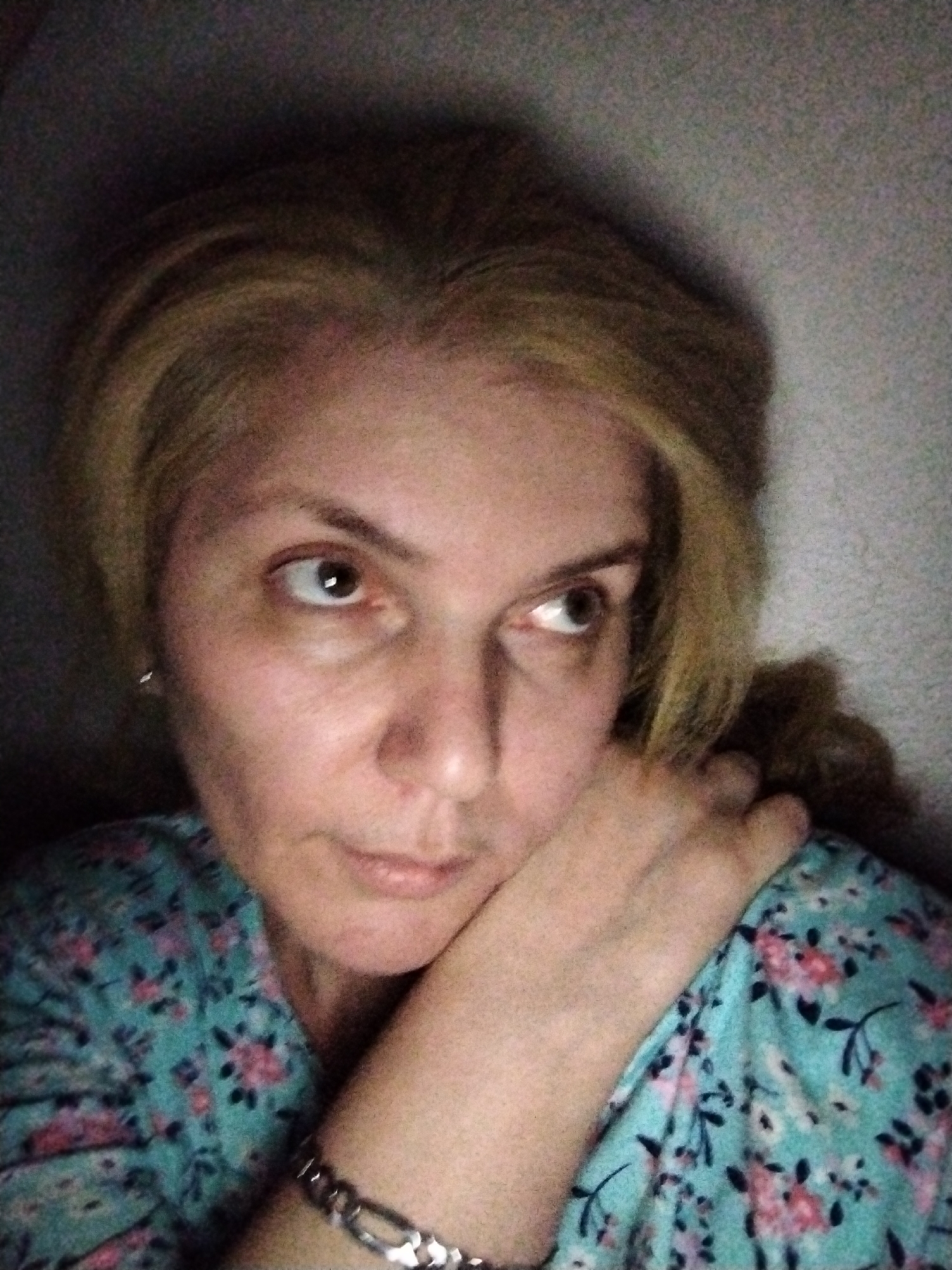 A woman with blonde hair is captured in a close-up, selfie-style photograph, her face tilted slightly upwards and to the right. She is resting her chin on her right hand, which is positioned under her jaw. Her expression appears pensive, with her eyes looking off into the distance. She is wearing a teal-colored blouse adorned with a floral pattern of pink and black flowers. A silver bracelet is visible on her left wrist. The background is a textured wall, dimly lit, with shadows creating a somber atmosphere. The scene is indoors, with the lighting suggesting it could be nighttime or a dimly lit room. No specific location in Al Mas'oudi, United Arab Emirates can be determined from the visual information provided.
