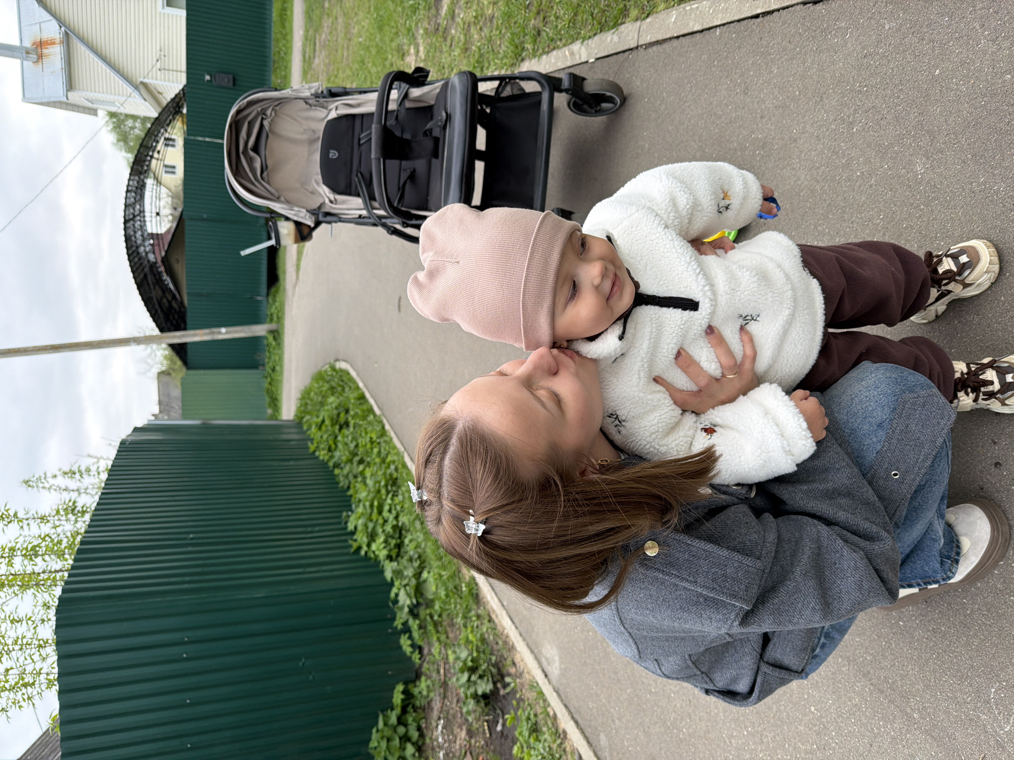 A woman is crouching on an asphalt surface, embracing a baby wearing a pink hat and a white, fleece-like jacket with brown pants and patterned sneakers. The baby is facing the woman and has a slight smile. The woman is wearing a gray jacket, jeans, and white shoes, and she has butterfly clips in her brown hair. Behind them is a black stroller. To the left is a green metal fence with a strip of greenery along the base, and beyond the fence is a building with a dark structure. The image was taken in Reshetnikovo, Russia.
