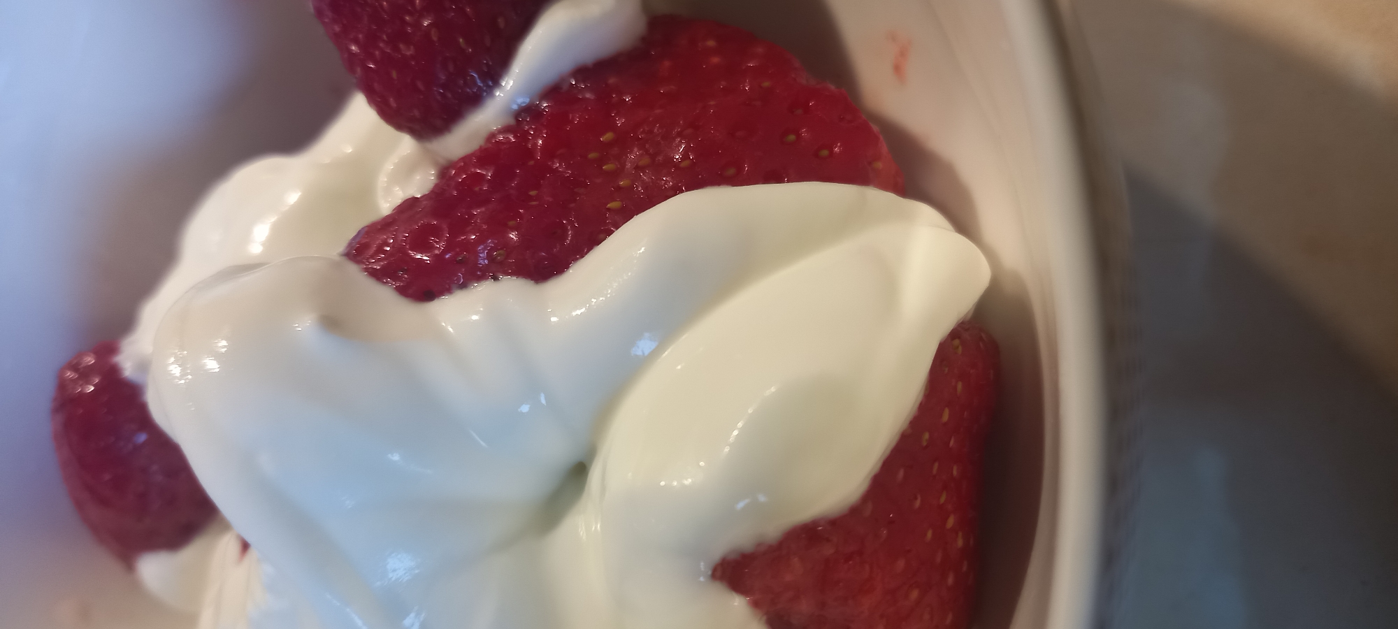 A close-up shot captures a bowl of strawberries topped with whipped cream. The strawberries are a vibrant red, with visible seeds. The whipped cream is thick and white, with soft folds and a glossy sheen. The scene appears to be indoors, with diffused lighting. There is no indication of the location in Kryvyi Rih, Ukraine.
