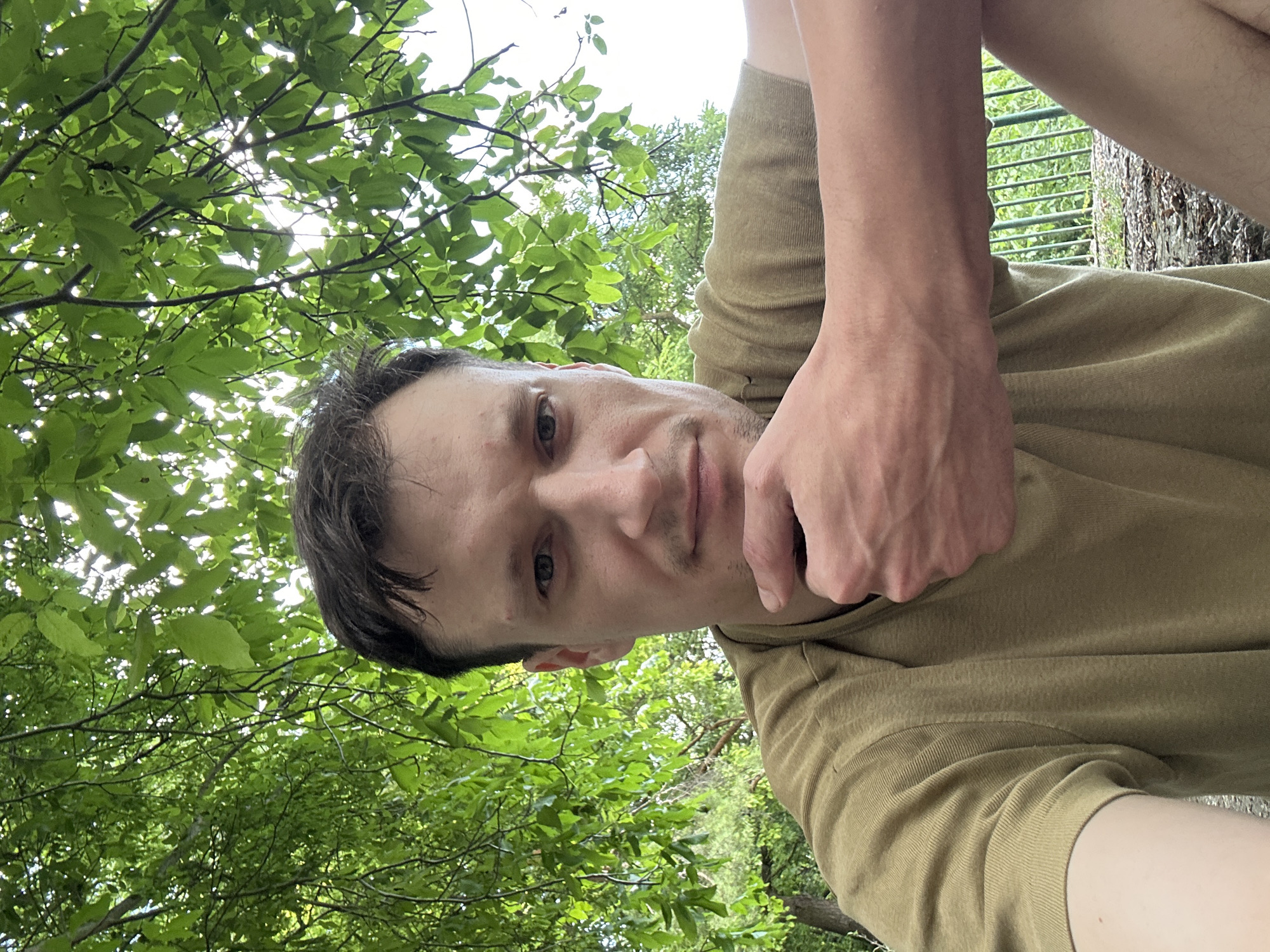A man is sitting outside under trees, holding his chin with his left hand. He is wearing a light brown t-shirt. The background is filled with green leaves and branches, with a hint of sky visible. The ground appears to be covered in dirt and possibly some leaves. The image is taken from a low angle, looking up at the man. The location is identified as Makiv, Ukraine.