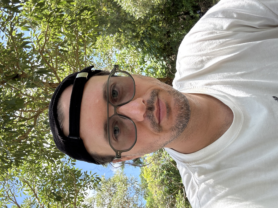 A man takes a selfie under trees, looking up at the camera. He wears a white t-shirt, a black hat with a strap, and metal-framed glasses. The background is filled with green foliage and glimpses of the sky. The man has short hair, light eyes, and a trimmed beard. The image is brightly lit with natural sunlight.