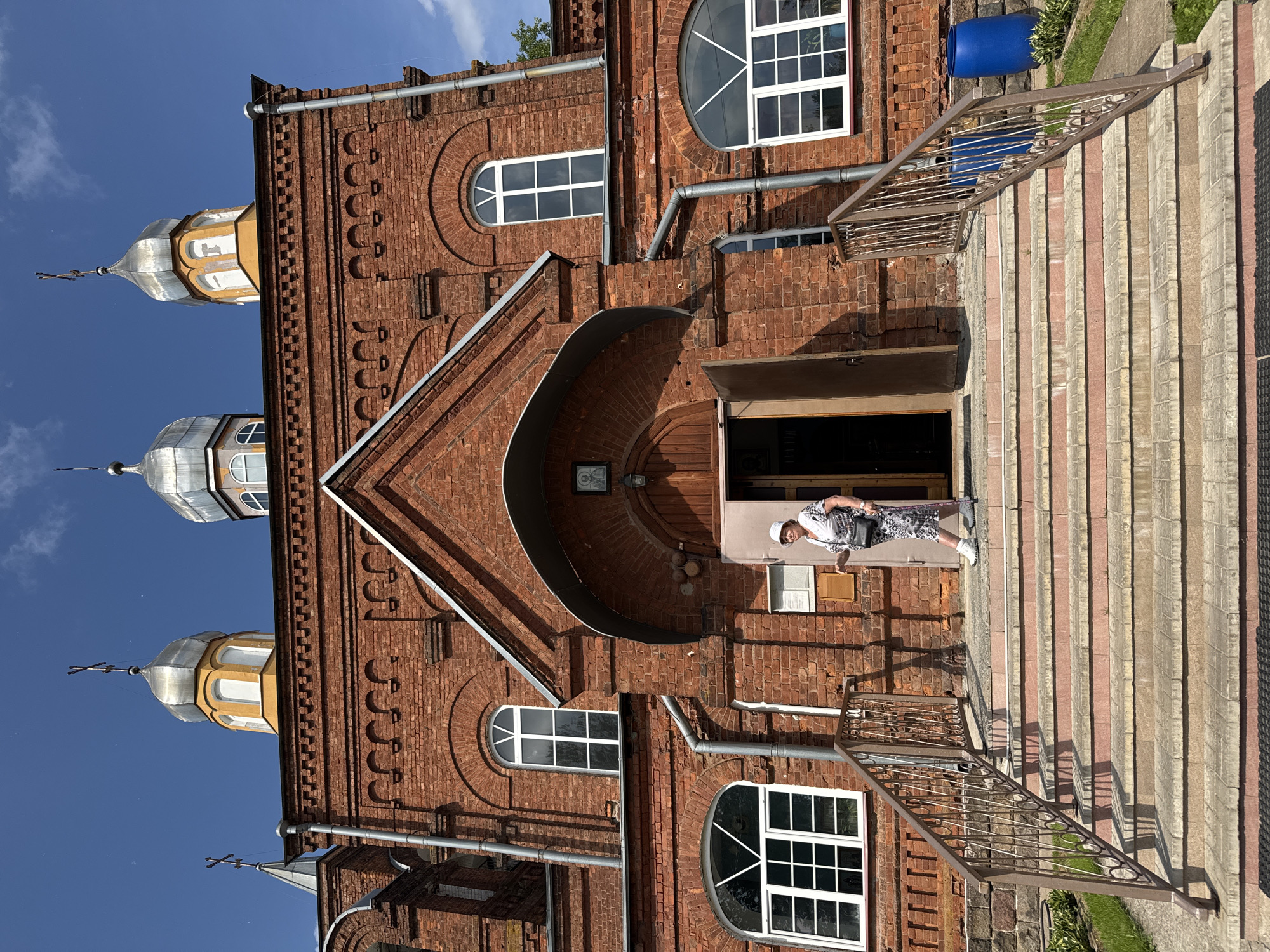 An elderly woman in a white hat and a patterned dress stands on the steps of a red brick church in Chudovo, Russia. She holds a walking stick in her right hand. The church features arched windows, a prominent entrance with a wooden door, and three onion domes topped with crosses against a clear blue sky. A metal railing lines the stairs, and two large blue barrels are visible to the right of the church. The scene is illuminated by bright sunlight, casting shadows on the steps.
