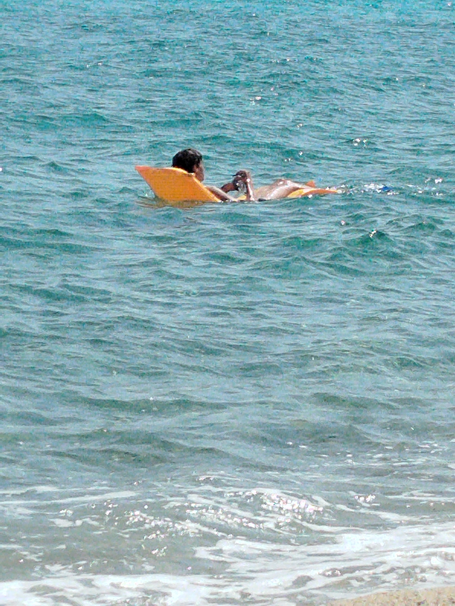 Two people are floating on a yellow raft in the sea at Sithonia Municipal Unit, Greece. One person, partially visible from the back, is lying on their stomach, propped up on their elbows. Another person is partially submerged next to them, appearing to be floating on their back. The water is a clear blue-green with visible waves and reflections of sunlight. The raft is yellow and buoyant. The background is a vast expanse of blue water. The scene is peaceful and appears to be a recreational activity in a coastal area.