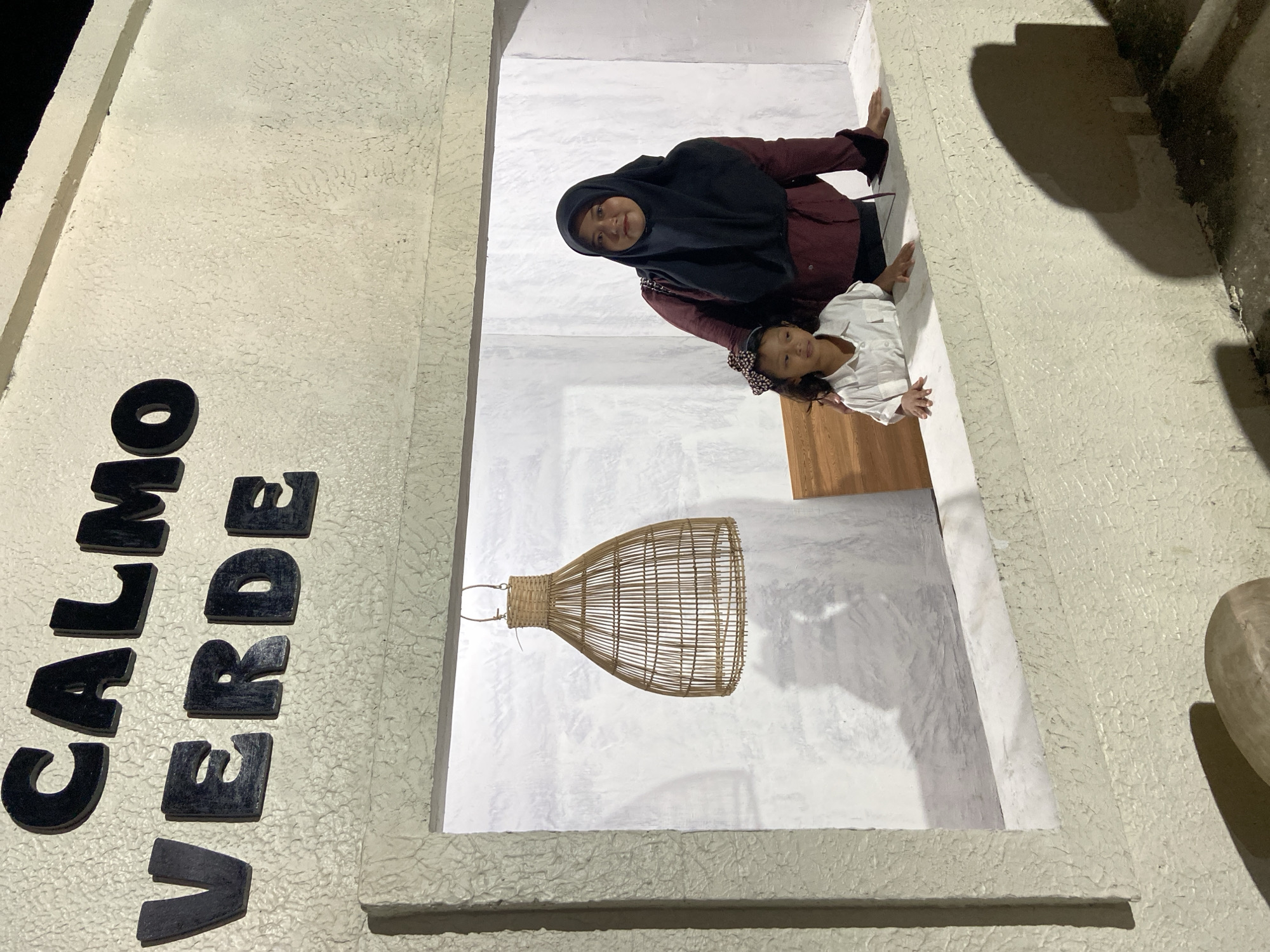 Two individuals, an adult and a child, are positioned in an opening or archway. The adult, wearing a dark hijab and a maroon jacket, is looking towards the viewer with a slight smile. To her right, a child in a white shirt smiles and looks forward. They appear to be posing for a photograph.
The setting is indoors, with white walls and a textured, light-colored exterior wall visible. A woven basket hanging from the ceiling is prominent in the background. To the left of the frame, large black letters are attached to the exterior wall, spelling out "CALMO VERDE". The lighting suggests it is nighttime or dimly lit, and the shadows cast indicate artificial lighting. The image was taken in Kesiman, Indonesia.