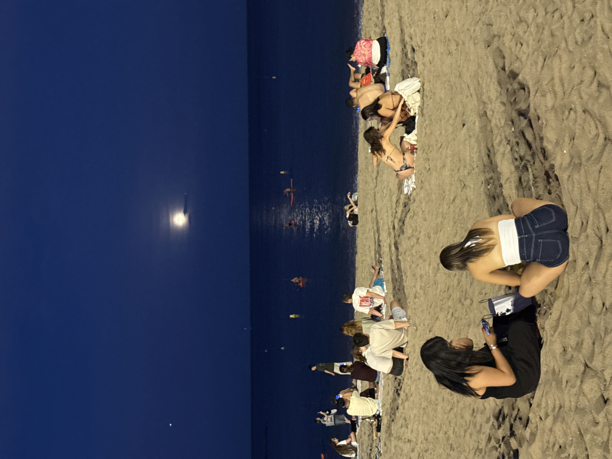 The image depicts a nighttime scene on a sandy beach, likely in Barcelona, Spain, given the presence of a clear night sky with a full moon and a distant bright star or planet. The sea is calm and dark blue, reflecting the moonlight. Several groups of people are scattered across the beach, some sitting on blankets and others lying down. In the foreground, two women are seated on the sand, one in a black dress looking at her phone, and the other in a white top and denim shorts facing away from the camera. Further back, various groups are seen socializing and relaxing, with some individuals in swimwear. In the water, a few people are visible on paddleboards or small boats. The overall atmosphere is one of casual evening leisure.