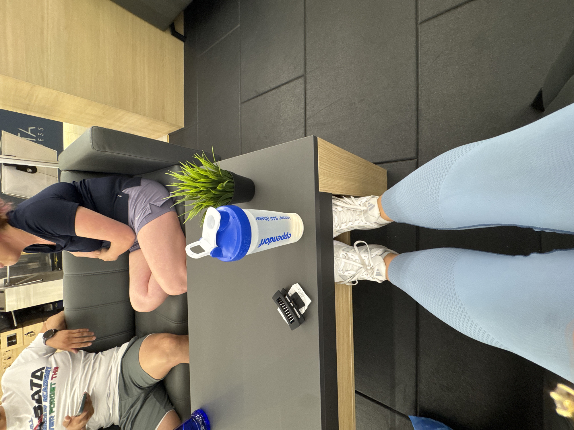 The image, oriented correctly, displays an indoor waiting area or lounge, likely within a fitness center in Sofia, Bulgaria. Two individuals are seated on a dark grey sofa. To the left, a person, possibly female, wears a dark blue top and grey shorts, with legs crossed, appearing to rest. To the right, a person, possibly male, wears a white t-shirt with text and grey shorts, looking down at a smartphone held in their hands. They wear a dark watch on their left wrist.
In the foreground, a person's legs and feet are visible, wearing light blue, textured athletic leggings and white Adidas sneakers. These legs are positioned near a dark grey rectangular table with wooden accents. On the table, a blue and white shaker bottle is placed, featuring the visible text "Eppendorf" and "Innova S44i Shaker". A small potted artificial green plant sits on the table, along with a black hair clip and a small white paper or tag.
The flooring consists of dark grey square tiles. In the background, there's a light wooden wall and a partial view of a sign with the letters "FITNESS" and "ESS" visible, further suggesting a gym or workout facility setting. The overall atmosphere appears relaxed, with individuals resting after or before a workout.
