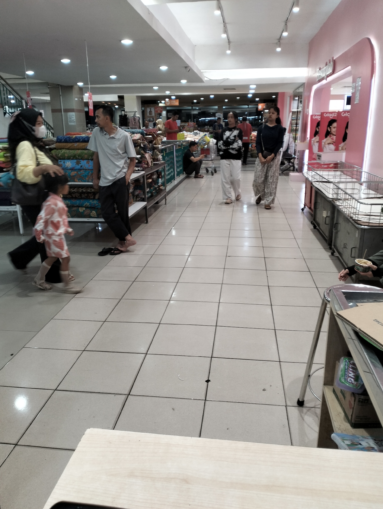 The image depicts a retail environment, possibly a department store, in Ungaran, Indonesia. In the foreground, a young child in a pink and white patterned outfit is walking, accompanied by an adult wearing a yellow top and a black headscarf. To their left, a man in a light grey polo shirt and black trousers is standing. Further into the background, other shoppers are visible. On the right side of the image, a pink counter or display area with signage featuring images of women is present, along with metal shopping carts. The floor is tiled in a grid pattern, and fluorescent lighting illuminates the space. The overall scene suggests a busy commercial setting.