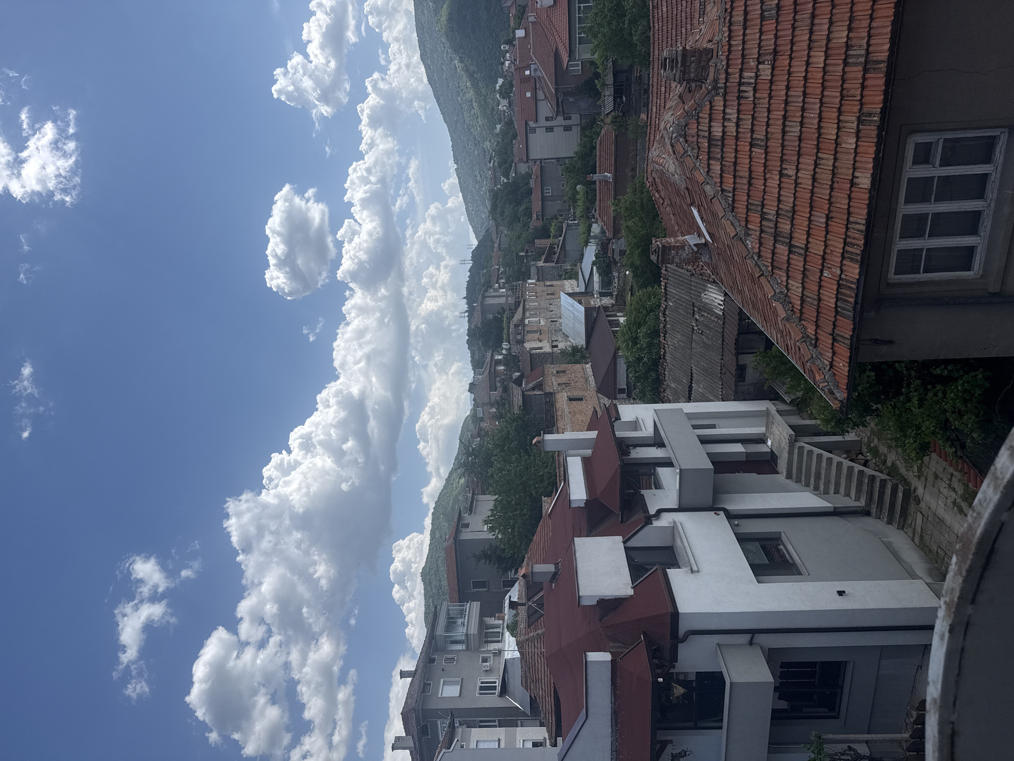 View from a high vantage point showing houses and buildings with red tile roofs in a city in Unknown. A range of mountains is in the distance behind the city. The sky is light blue and has large white cumulus clouds.