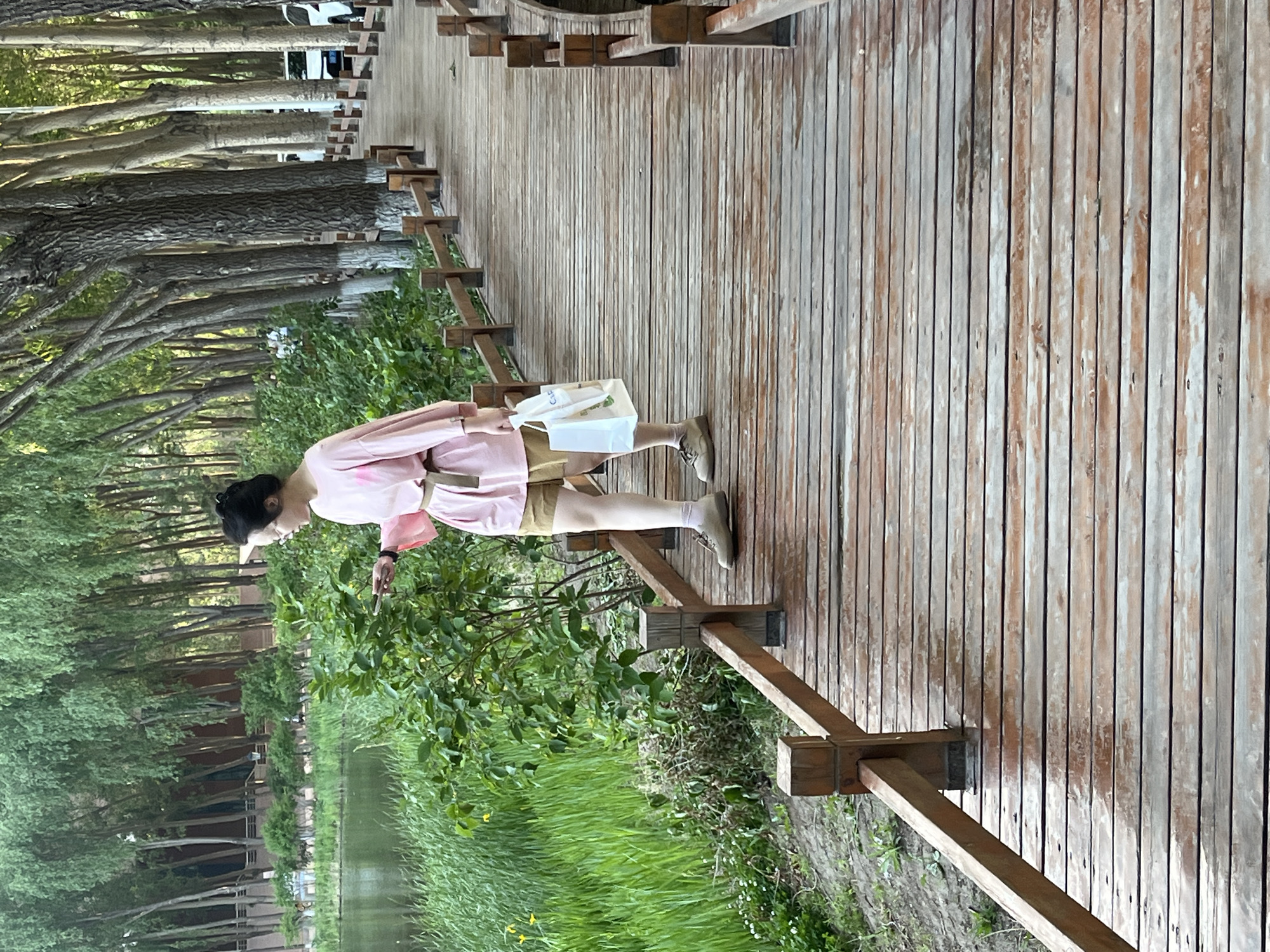 A woman stands on a wooden walkway surrounded by trees and lush greenery, seemingly in a park or nature preserve. The walkway has a wooden railing. She is wearing a light pink shirt and khaki shorts, with white socks and brown shoes. She is holding something small in her hand near the greenery. The environment includes a body of water, likely a river or pond, with tall grasses along the banks. The trees form a dense canopy in the background. Based on the request, this image is likely taken in Changchun City, China.