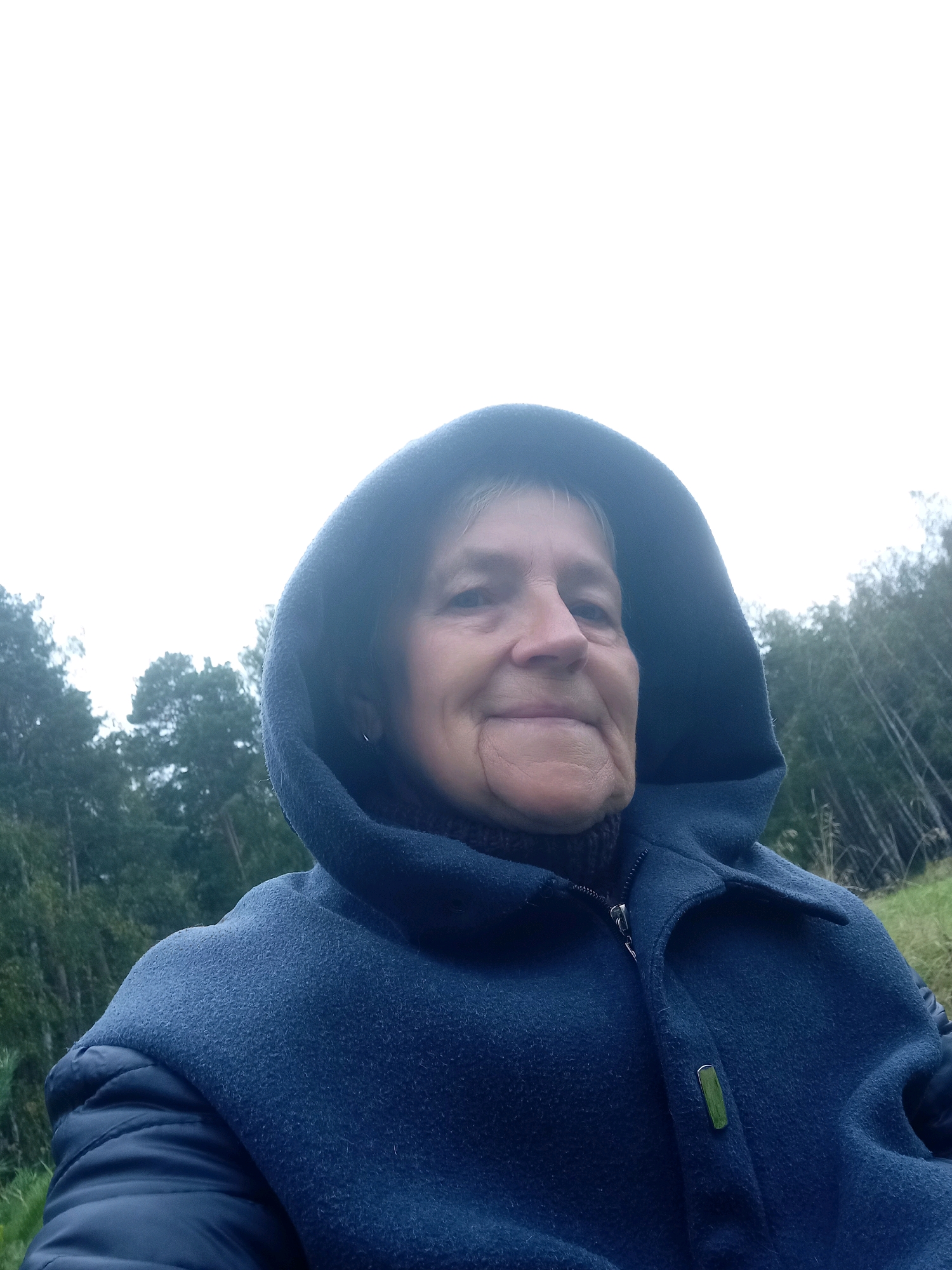 A close-up, low-angle selfie shows a woman with light-colored hair wearing a dark blue hooded coat. She is looking directly at the camera with a slight smile. Her face is illuminated from above by the overcast sky. The coat's hood is up, framing her face. In the background, a dense forest of trees with thin trunks is visible under a bright, featureless sky, suggesting an outdoor setting in a natural environment, possibly within the city of Degtyarsk, Russia, characterized by its surrounding wooded areas and industrial landscape.