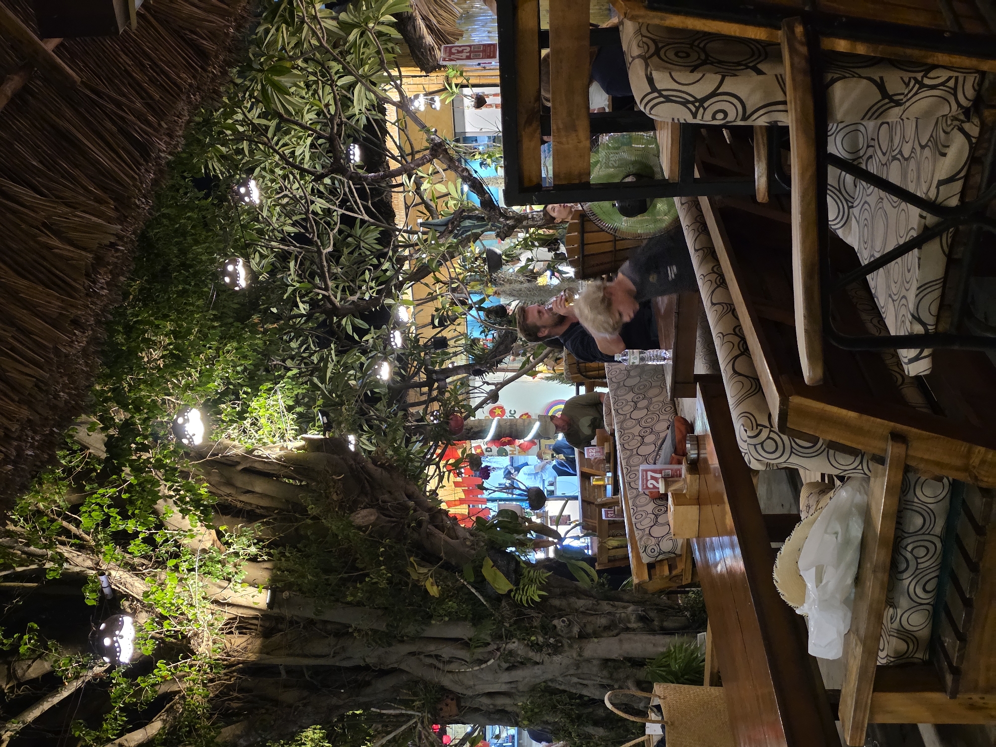 This image depicts an evening scene at an outdoor or semi-outdoor restaurant in Mũi Né, Vietnam, characterized by a lively, tropical ambiance. The setting is dominated by large trees and dense green foliage that are integrated into the dining area, illuminated by numerous artificial lights.
Guests are seated at sturdy wooden tables and chairs, the latter featuring cushions with a distinctive black and white concentric circle pattern. In the central foreground, a bearded man is holding a bottle, possibly drinking, with another individual whose blond hair is visible next to him. A green electric fan is placed on an adjacent table, suggesting warm weather.
The lighting is a notable feature, with numerous small, spherical lights strung throughout the trees, creating an ambient glow. Additionally, larger, decorative lights resembling yin-yang symbols emit bright white light, highlighting sections of the tree trunks and branches. On the left, a traditional thatched roof is visible, adding to the rustic, local character.
In the background, red lanterns contribute to the vibrant decor, and a person is faintly visible wearing a conical hat (nón lá), a strong cultural indicator of Vietnam. On a table in the foreground, a red stand displays the number "27". Another partially obscured sign in the far background appears to have the text "NHAN" or "THANH MAI". A straw hat and a white plastic bag hang from one of the chairs, suggesting personal belongings. The overall scene conveys a relaxed, social dining experience amidst lush, illuminated greenery.