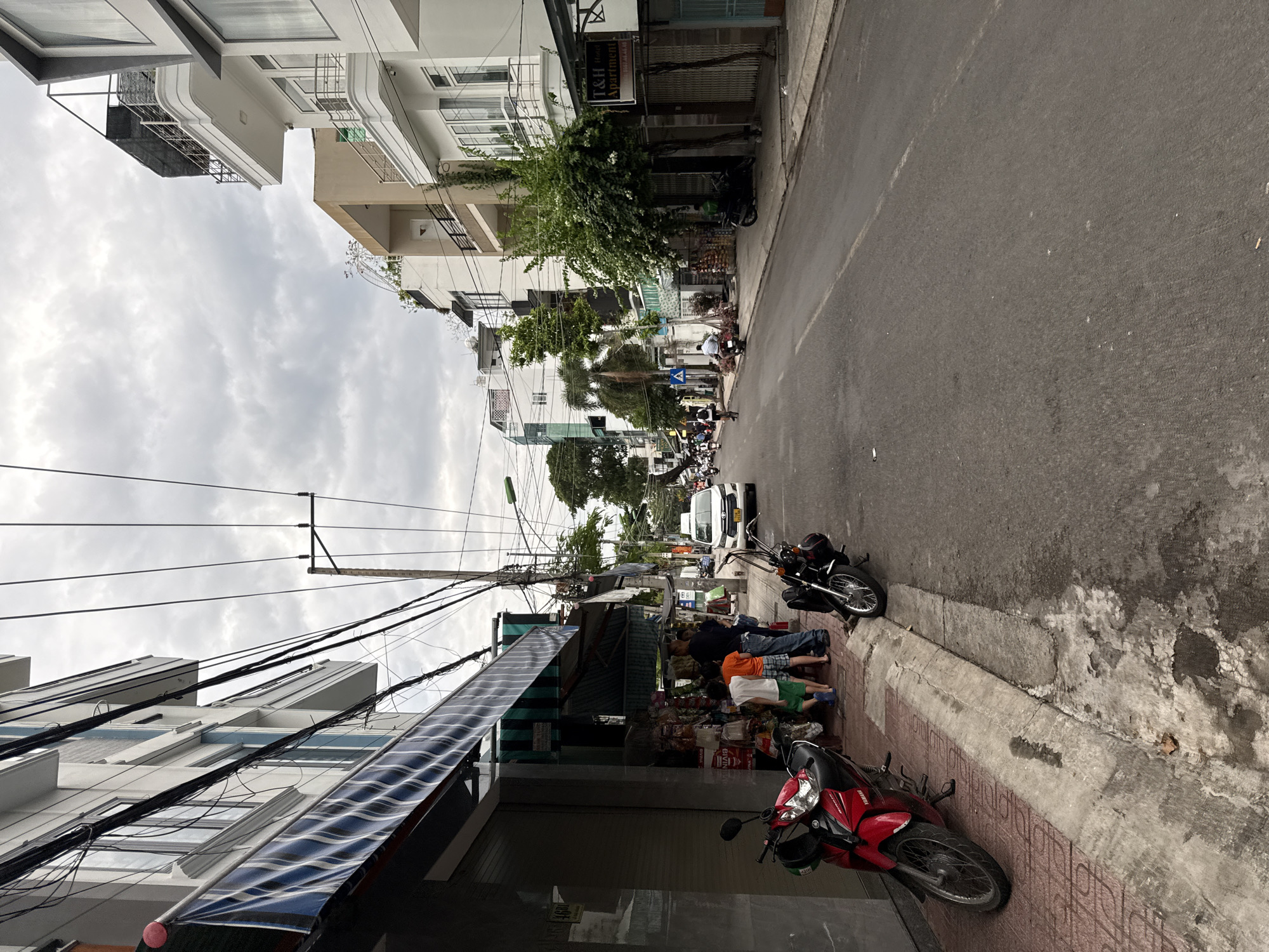 The image depicts a street scene in Nha Trang, Vietnam, likely on a cloudy day. The street is lined with multi-story buildings, many of which appear to be apartments or small businesses. Overhead, a dense network of electrical wires is prominently visible.
On the left side of the street, a red Yamaha motorcycle is parked on a tiled sidewalk. Adjacent to it, a store with a rolled-down metal shutter displays various items on shelves outside. A man and three children are standing near the store, with the children appearing to browse the merchandise. Another motorcycle, black and parked to the right of the first, is partially obscured by the children and the store display.
The street itself is paved and appears to be moderately busy, with several vehicles visible in the distance, including a white car and multiple motorbikes. People are seen riding motorbikes and bicycles on the road. The architecture of the buildings is varied, with some featuring balconies and modern designs, while others have awnings and more traditional shop fronts. Trees and foliage are present along the street, indicating a somewhat urbanized but green environment. A sign for "T&H Hotel APARTMENT" is visible on a building further down the street. The overall impression is of a typical street in a Vietnamese city, characterized by its narrow streets, dense infrastructure, and active street life.