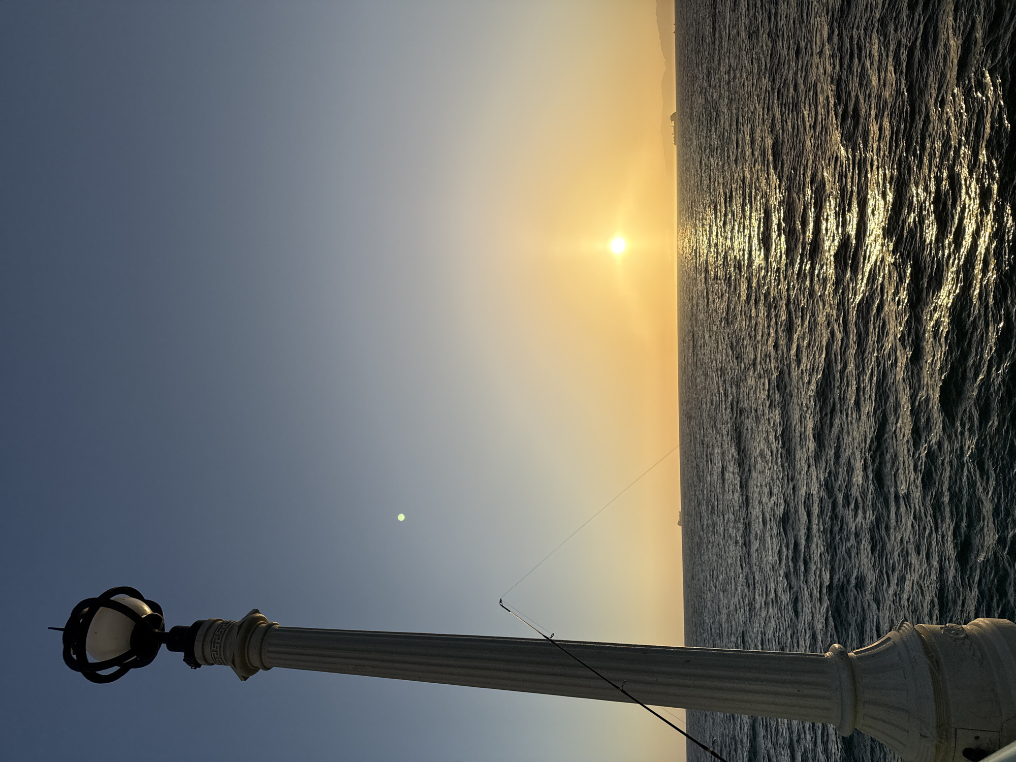 This image depicts a sunset over the Pacific Ocean, viewed from the Manhattan Beach Pier in Manhattan Beach, California, United States of America. In the foreground, a white lamppost with a decorative base stands to the left, its shadow cast towards the right. A fishing rod is propped against the lamppost, with its line extending towards the ocean. The sun is setting on the horizon, casting a warm golden glow across the water and the sky. The ocean is relatively calm with gentle waves, and several ships are visible in the distance. The sky transitions from a clear blue at the top to a soft orange hue near the horizon.