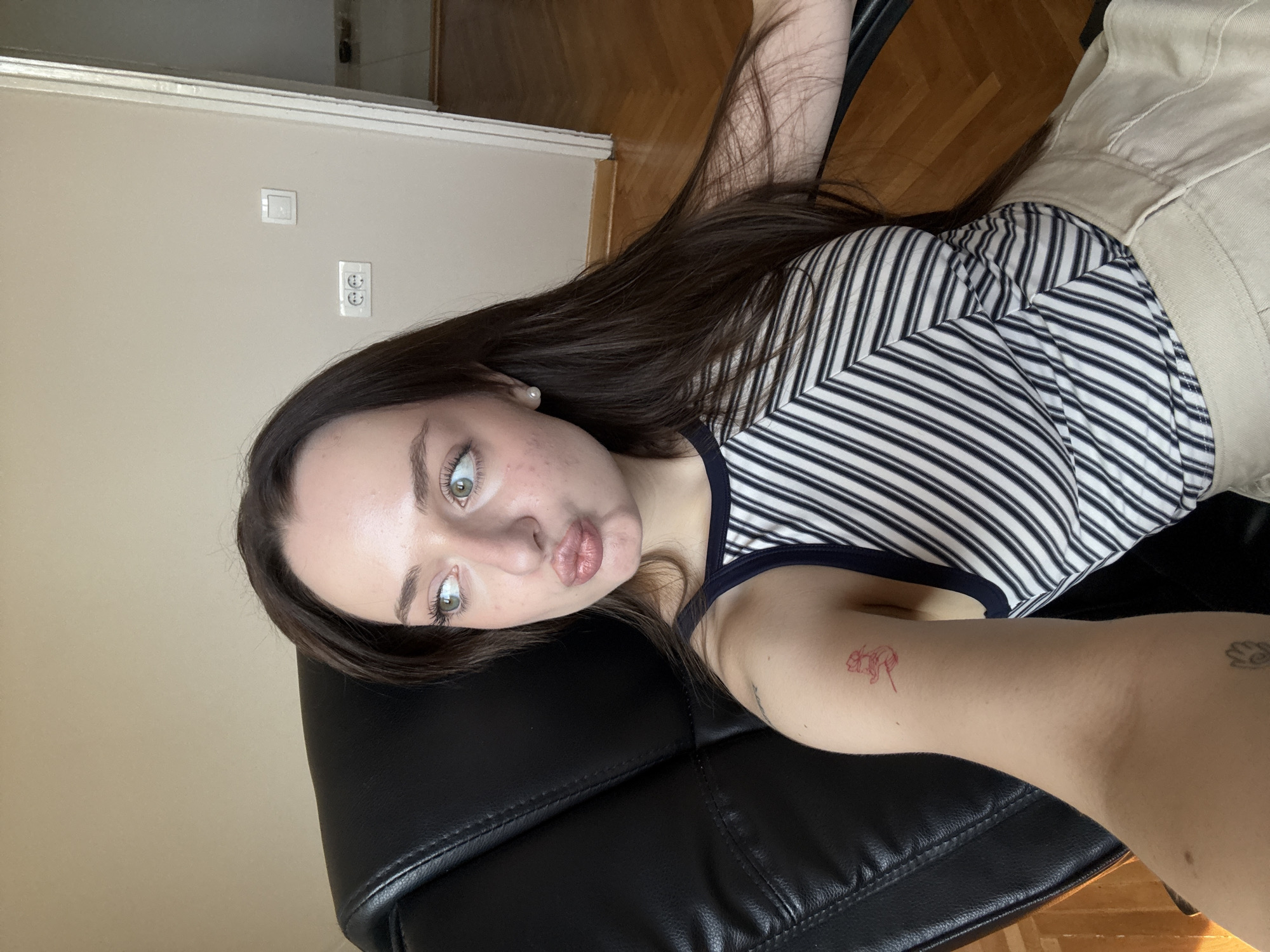 A young woman with long, dark hair and fair skin, wearing a striped tank top and light-colored pants, sits in a black office chair. She is looking to her right, her lips pursed in a playful expression. She has a small, red tattoo of a flower on her upper left arm, and another faint tattoo on her forearm. The background shows a beige wall with two electrical outlets and a light switch, and a doorway leading to another room with wooden flooring. The scene appears to be indoors, likely in a residential or office setting.