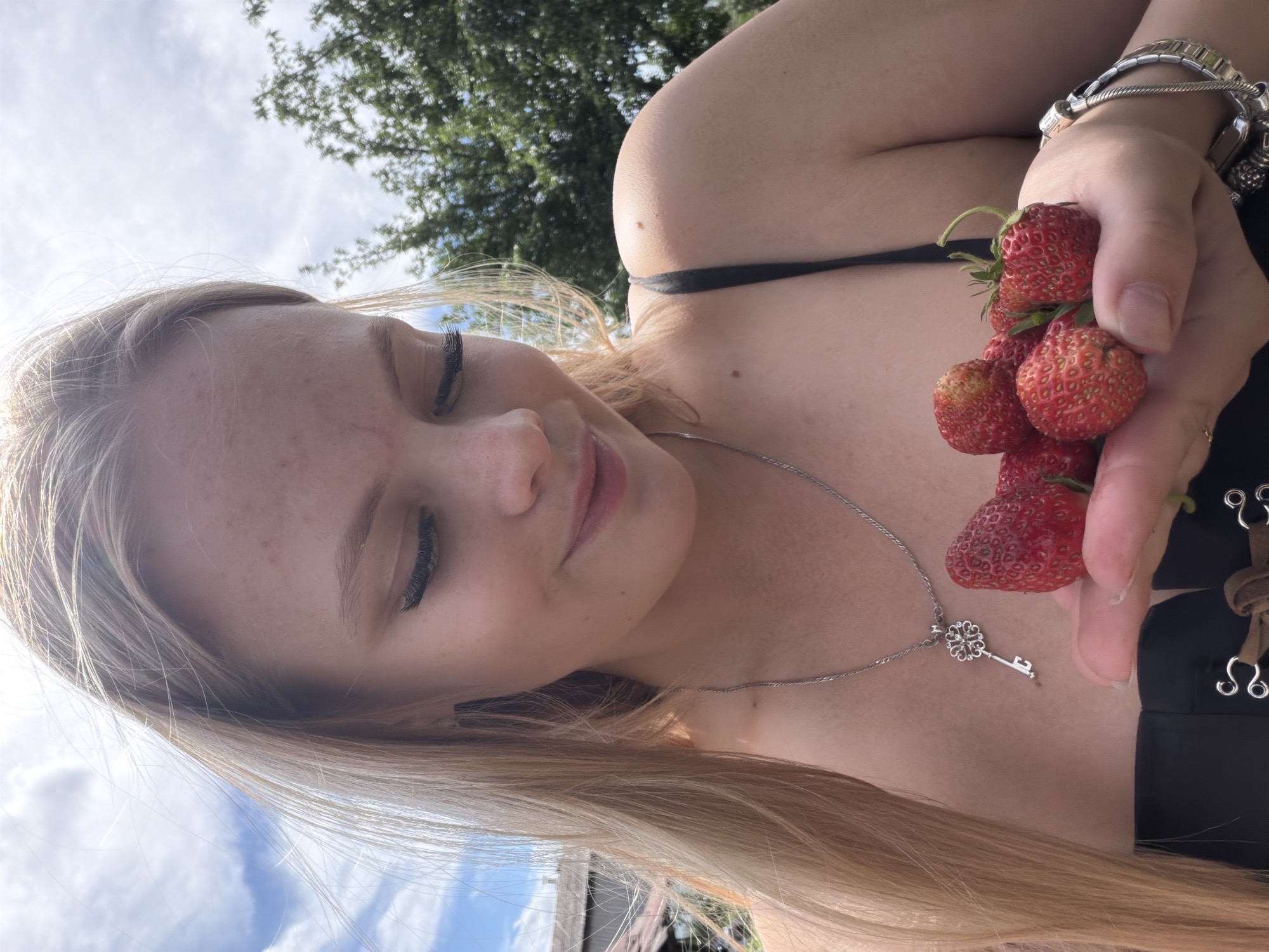 In an outdoor setting, a young woman with blonde hair and freckles holds a handful of strawberries. She is wearing a black top and a silver necklace with a key pendant. The background consists of trees, a building, and a cloudy sky. The image appears to have been taken during the daytime.