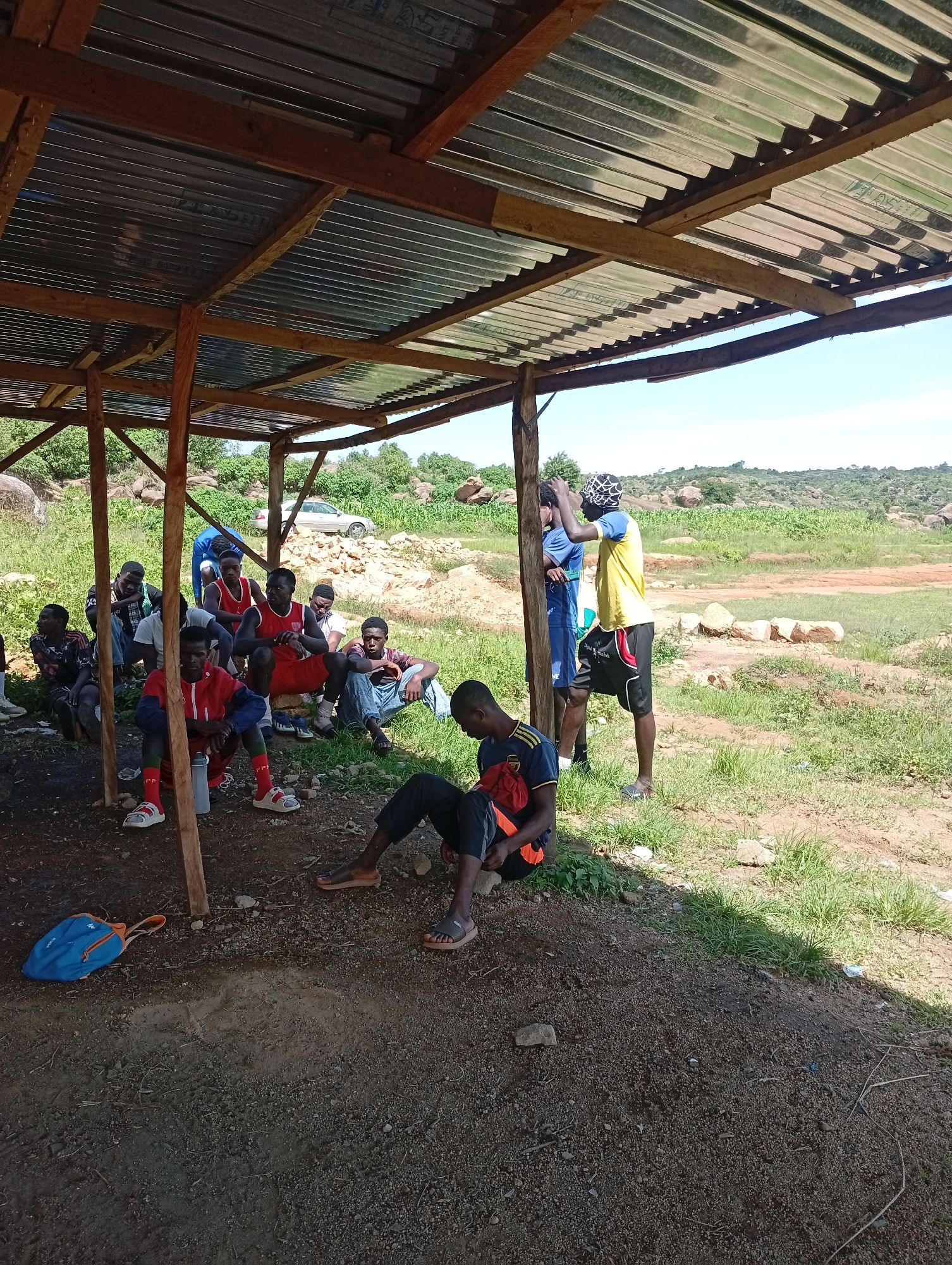 The image depicts an outdoor gathering of at least eleven young men under a makeshift shelter constructed with wooden posts and a corrugated metal roof. The shelter is situated in a dry, earthy area with sparse green grass and scattered rocks, typical of the landscape around Jos, Nigeria. The men are dressed in casual clothing, with some wearing athletic attire.
Several individuals are seated on the ground, some appearing to be resting or conversing. Two individuals stand towards the right side of the shelter, one wearing a yellow and black outfit, and the other in a blue shirt and shorts. A blue backpack lies on the ground near the left side of the shelter. In the background, a silver car is parked near a rocky, uneven terrain covered with vegetation, suggesting a rural or semi-rural setting. The overall scene appears to be a pause or a gathering point for a group of young men in a natural, somewhat undeveloped environment.