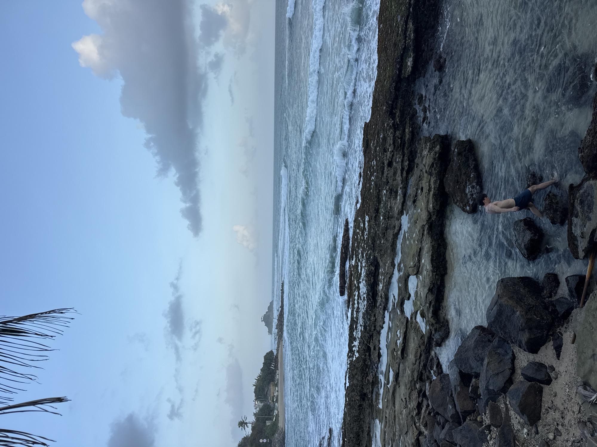 A young man, wearing dark swim shorts, walks through shallow water amidst rocks on the shoreline of Goviyapana, Sri Lanka. The man is positioned in the lower right quadrant of the frame, facing left and looking down. The sky above is a pale blue with scattered gray clouds. The ocean stretches to the horizon with moderate waves breaking onto the shore. The foreground consists of rough, dark rocks and some sand, with pools of water reflecting the sky. In the mid-ground, a sandy beach is visible with palm trees, structures, and small boats, leading to a rocky outcrop in the sea. The overall lighting suggests it is either early morning or late evening.