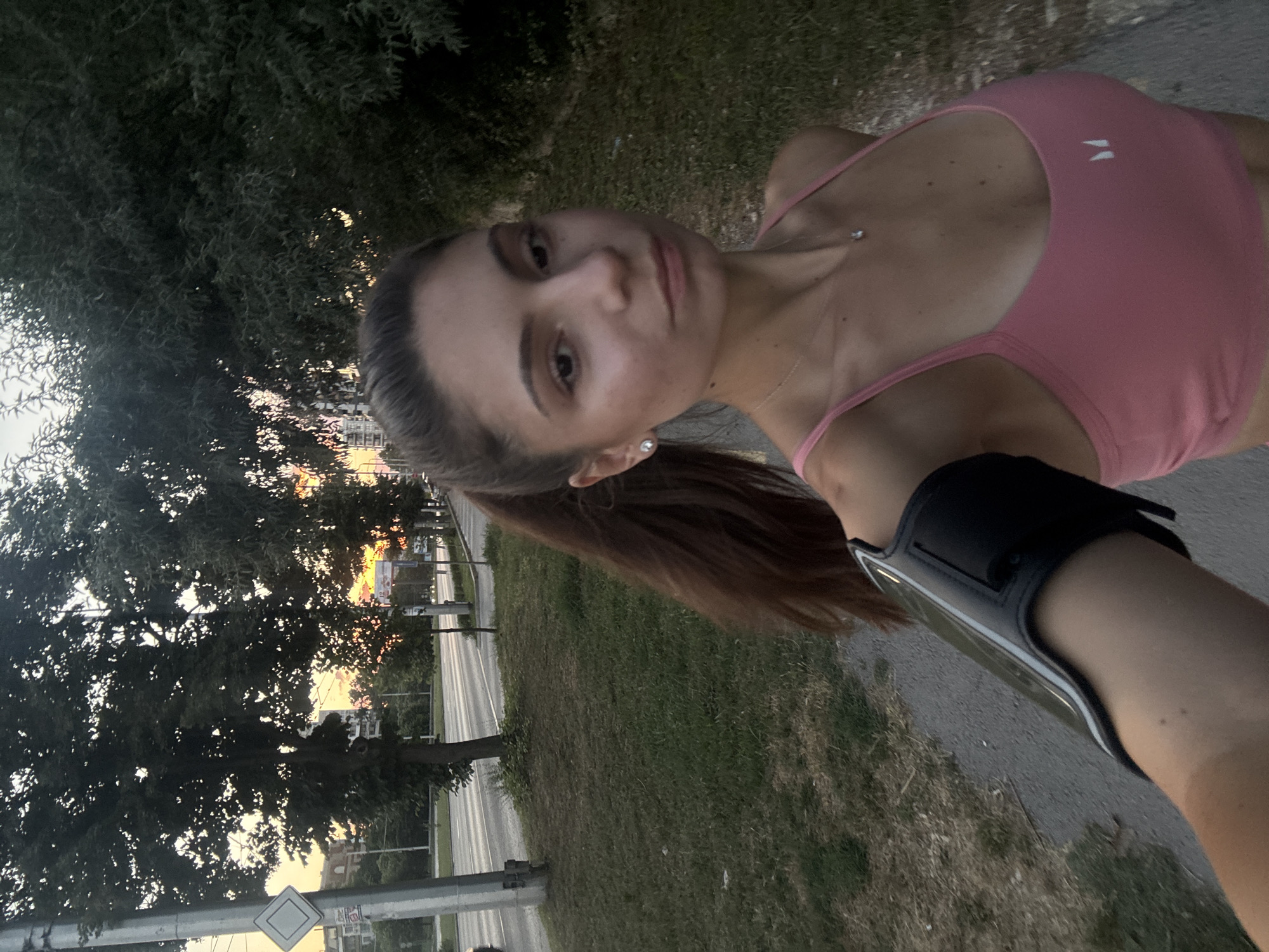A young woman with brown hair pulled into a ponytail is taking a selfie while jogging outdoors. She is wearing a pink sports bra and an armband on her left arm. The background shows a tree-lined sidewalk next to a road, with buildings and a sunset visible in the distance. The location is Varna, Bulgaria, possibly along the Varna-Sunny Beach highway.
