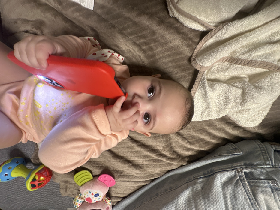 A baby in a peach-colored, star-patterned onesie lies on a brown blanket. The baby holds a red, angled object to their face, possibly chewing on it, with fingers near their mouth. Nearby are baby toys, including a colorful rattle and a plush toy. Partially visible are blue jeans and a cream-colored towel.