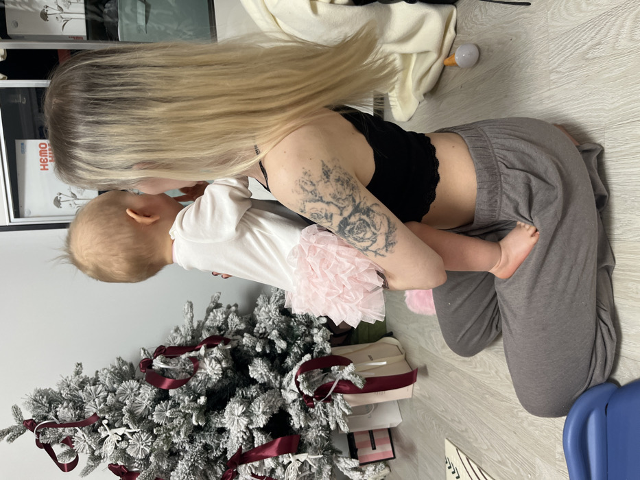 A woman kneels on the floor, embracing a baby dressed in a white onesie with a pink tutu. The woman has long blonde hair and a tattoo on her upper arm. A decorated Christmas tree is visible, along with gift bags. The woman is wearing a black top and gray pants. The scene appears to be indoors, likely a home setting.