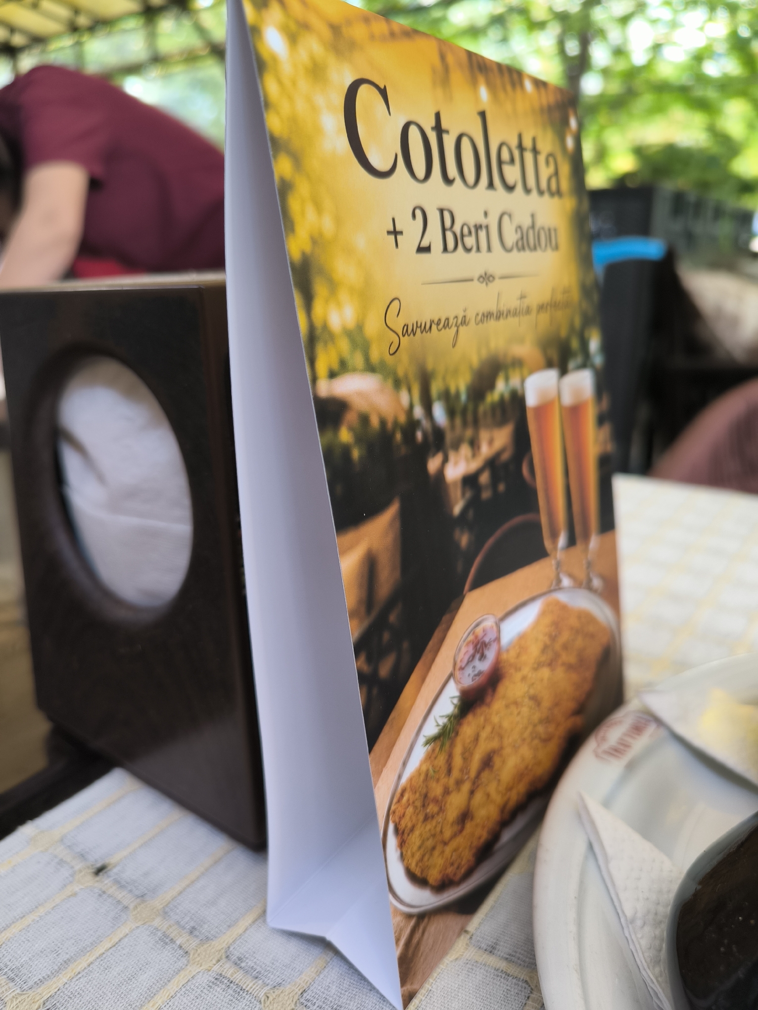 A close-up, eye-level shot shows a menu tent on a table outdoors, with a blurred background of trees and greenery. The menu tent is white and displays text and images. The text on the tent reads "Cotoletta + 2 Beri Cadou" and below that, "Savurează combinația perfectă". There is an image of a schnitzel with a side of sauce and rosemary, and two glasses of beer. To the left of the menu tent is a dark wooden tissue box with a circular opening revealing white tissues. In the background, a person wearing a dark red t-shirt is visible, their head and upper body blurred. The table is covered with a white and yellow checkered cloth. The scene appears to be in an outdoor seating area of a restaurant or cafe. The specific location in Chișinău, Moldova cannot be determined from the provided image.