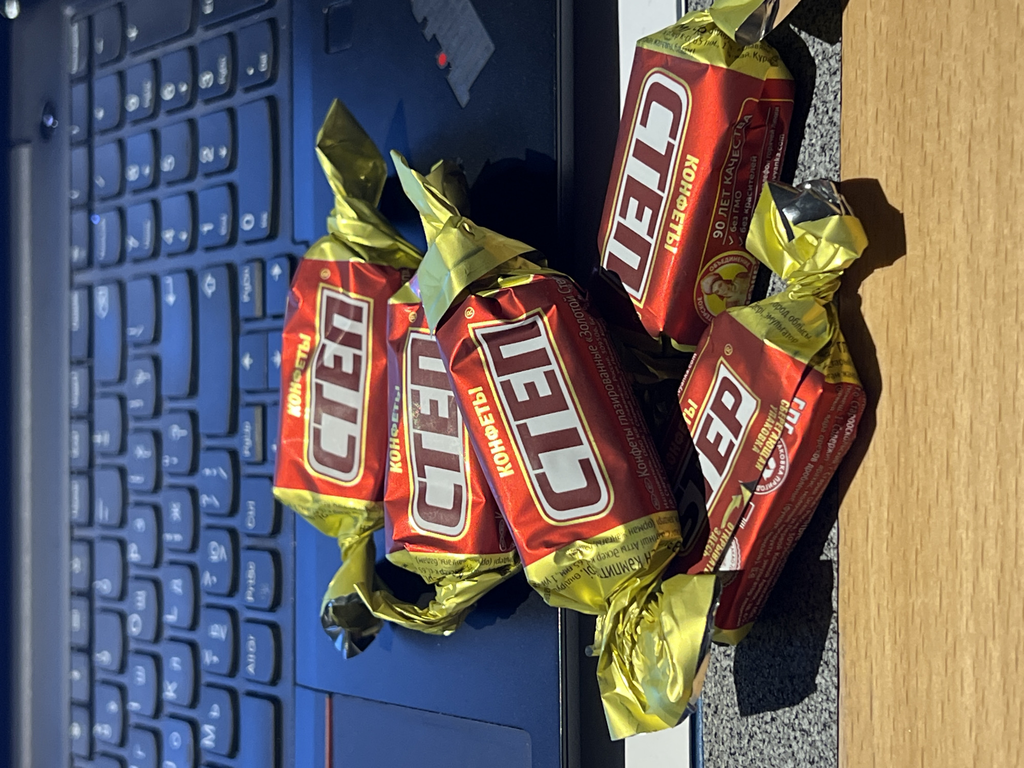 The image depicts five individually wrapped Step brand chocolates, likely of Russian origin due to the Cyrillic writing on the packaging, positioned on top of a laptop keyboard. The chocolates are wrapped in gold foil and have red and white packaging. The packaging includes information written in Russian, including the words "KONFETI" (candies), "STEP" (the brand name), and mention of "90 let kachestva" (90 years of quality). There is a reference to a manufacturer in the city of Stary Oskol, Russia and a city of origin Khimki, Russia.