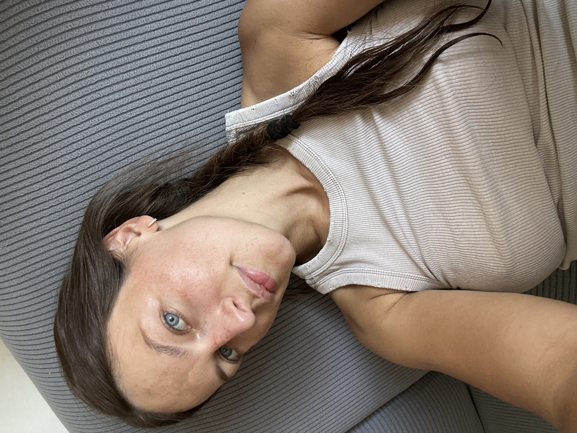 A person with brown hair and blue eyes lies on a gray ribbed surface, possibly a sofa or couch. They are wearing a light-colored, ribbed t-shirt. Their hair is pulled back with two black hair ties. The person's head is tilted, and they are looking directly at the camera with a neutral expression. The lighting suggests it could be daytime, and the setting appears to be indoors. There are no visible text elements in the image. The overall scene is casual and relaxed.