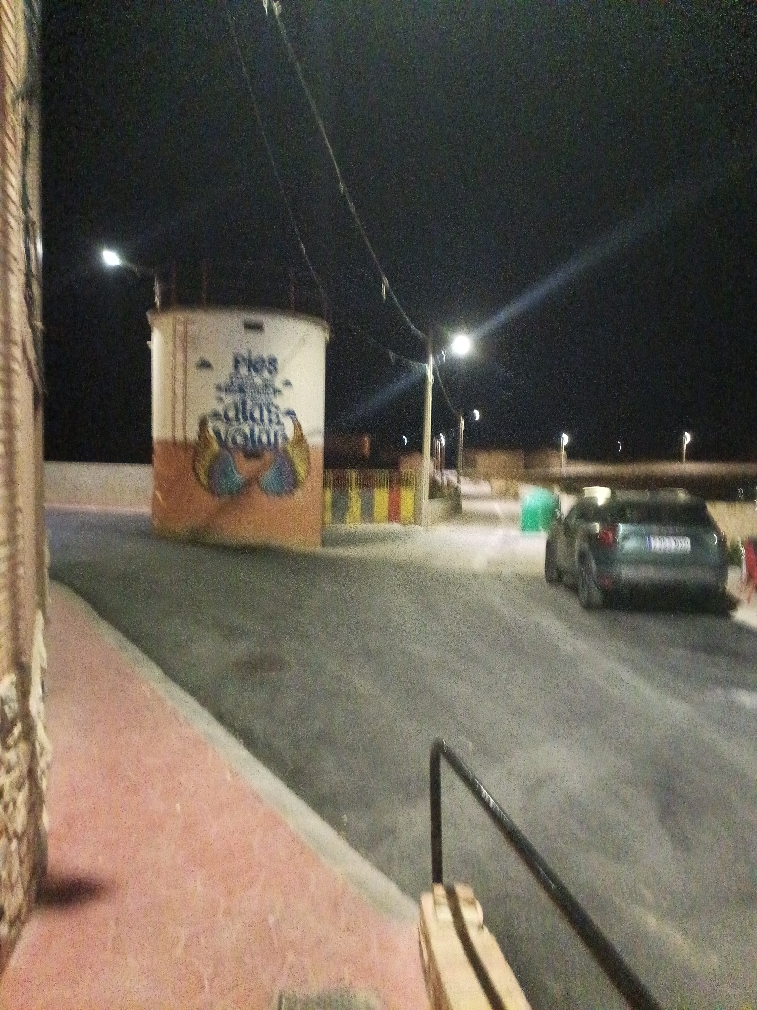 At night, on a street in Mainar, Spain, a cylindrical structure with faded text and artwork stands to the left of the road. The structure has a white top section with "Pies" and what appears to be stylized wings painted on its lower, orange section. The road is paved and curves to the right. A dark-colored car is parked on the right side of the road, facing away from the camera. Streetlights illuminate the scene, casting long shadows. A brick wall is visible on the far left of the image.