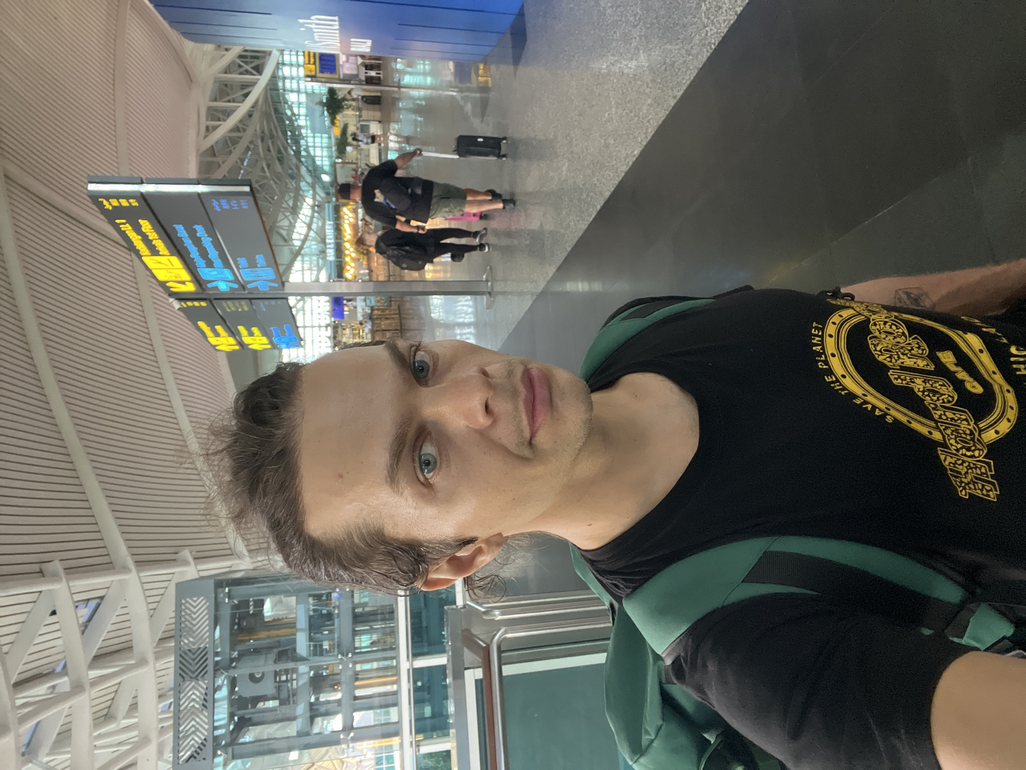 A man is taking a selfie in an airport. The airport has signs for "Kedatangan L1, Arrivals 1st Floor, Smoking Booth." There are people with luggage visible in the background. The man is wearing a black t-shirt with a "Save the Planet" design. The man appears to be at the Ngurah Rai International Airport in Kuta, Indonesia.
