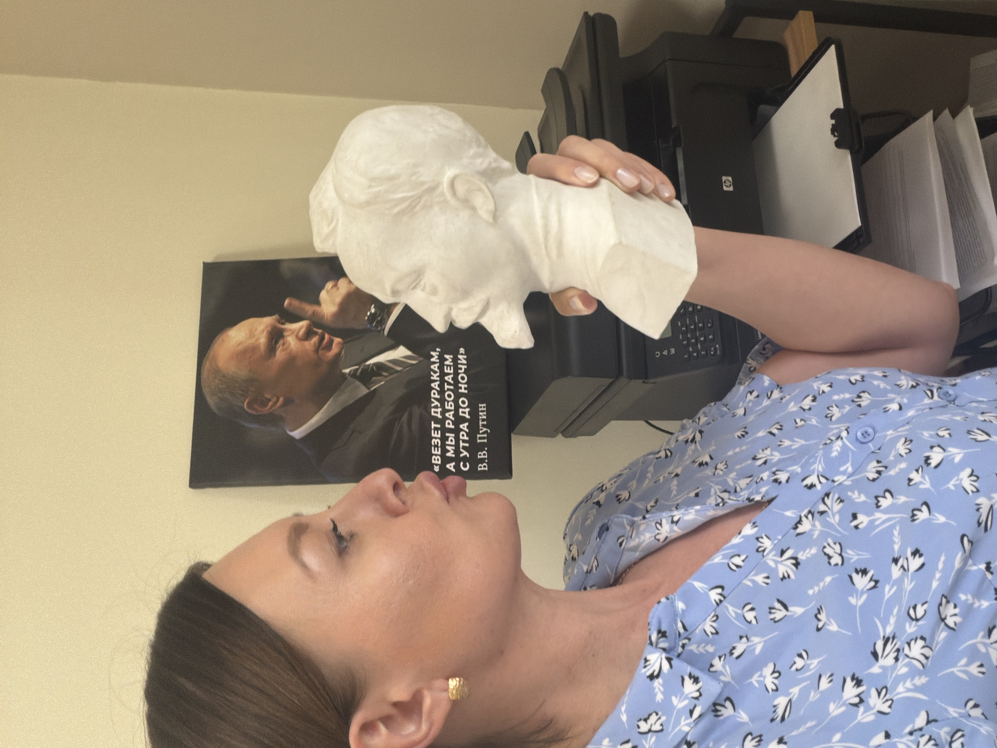 A woman is holding a white plaster bust of a man up to her face. In the background is a poster of Vladimir Putin with text written in Russian. The text translates to, "Fools get lucky, and we work from morning to night" and the text "V.V. Putin" below it. Next to the poster is a black printer and papers stacked on top of it. The woman is wearing a light blue floral shirt, and she has brown hair. She is wearing a gold earring.